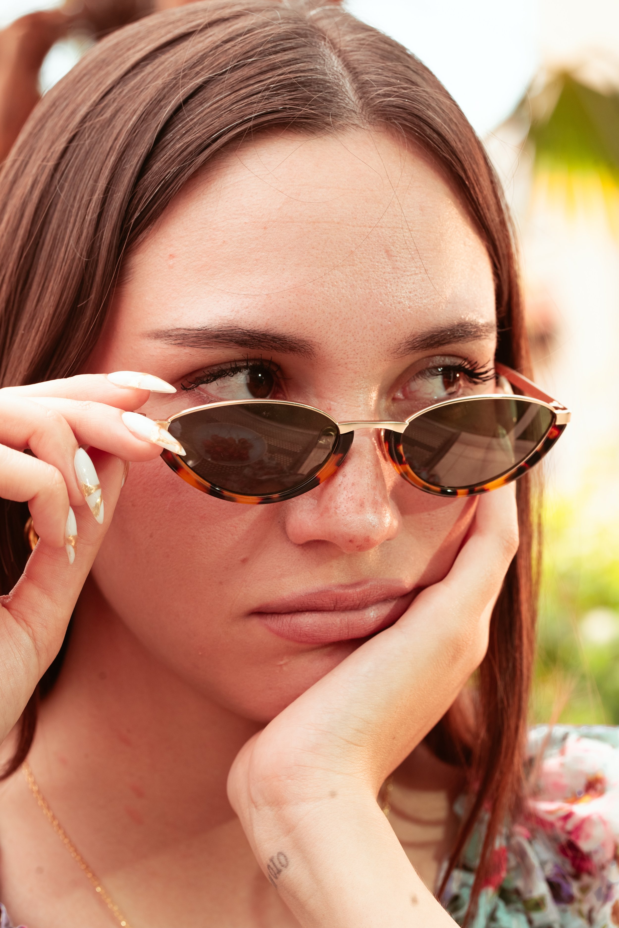 Ragazza con capelli castani, occhiali da sole tondi tartarugati, mani sul viso, sfondo di natura verde e luminosa.