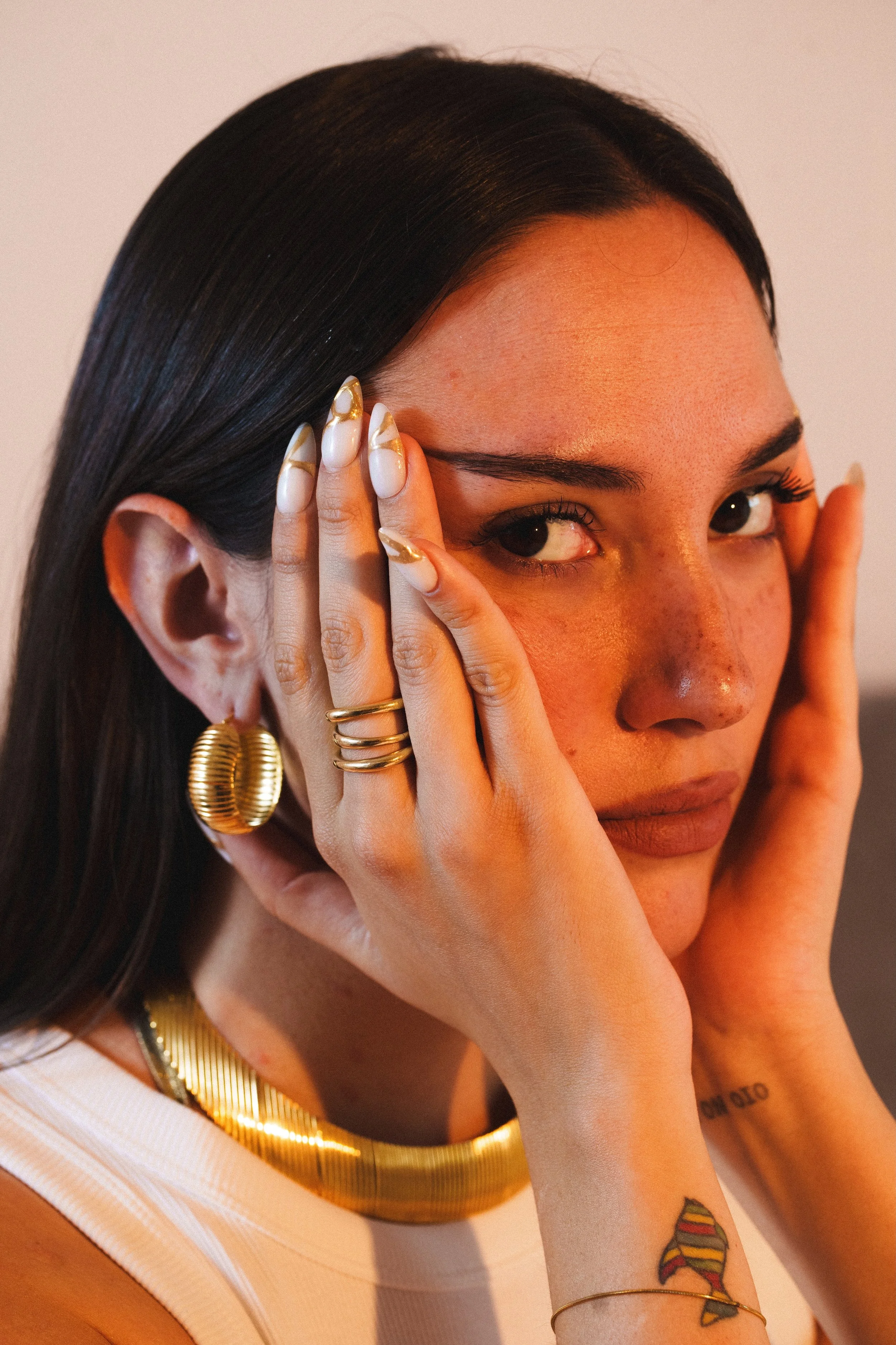 Ragazza con trucco naturale, orecchini grandi d'oro, anello, collana d'oro, tatuaggi sul braccio, posa con le mani sul volto che mette in risalto il viso.