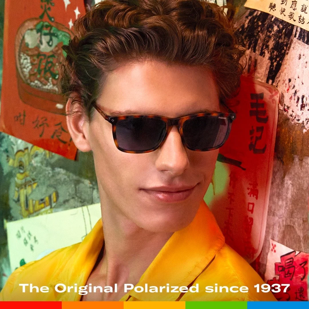 Ragazzo con capelli castani ricci, occhiali da sole tartarugati e giacca gialla, in un ambiente con decorazioni asiatiche, poster con scritte in cinese e sfondo colorato.