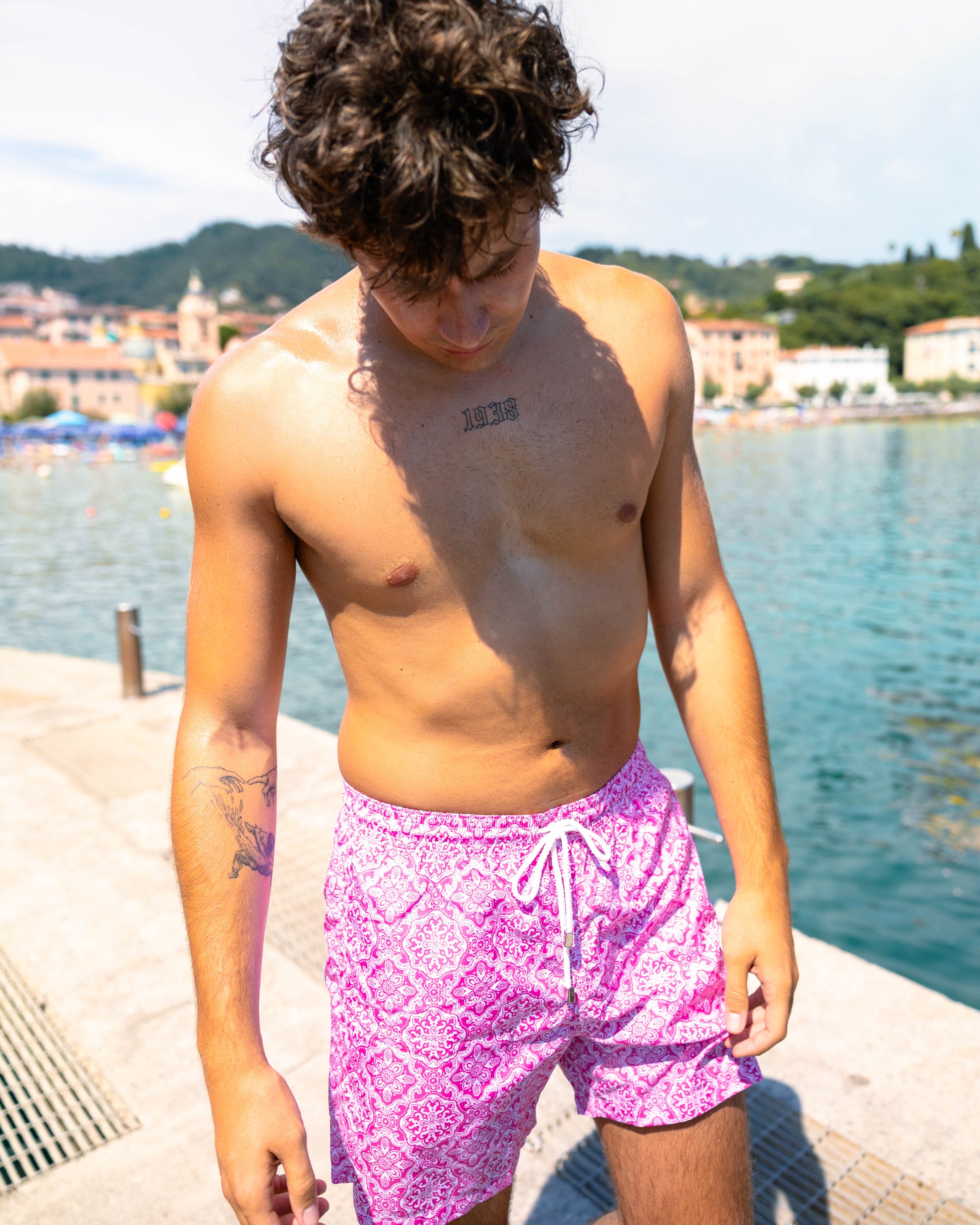 Ragazzo senza maglietta con tatuaggi, indossa pantaloncini rosa con disegni bianchi, si trova sul pontile affacciato su un lago o mare, con edifici e colline sullo sfondo.