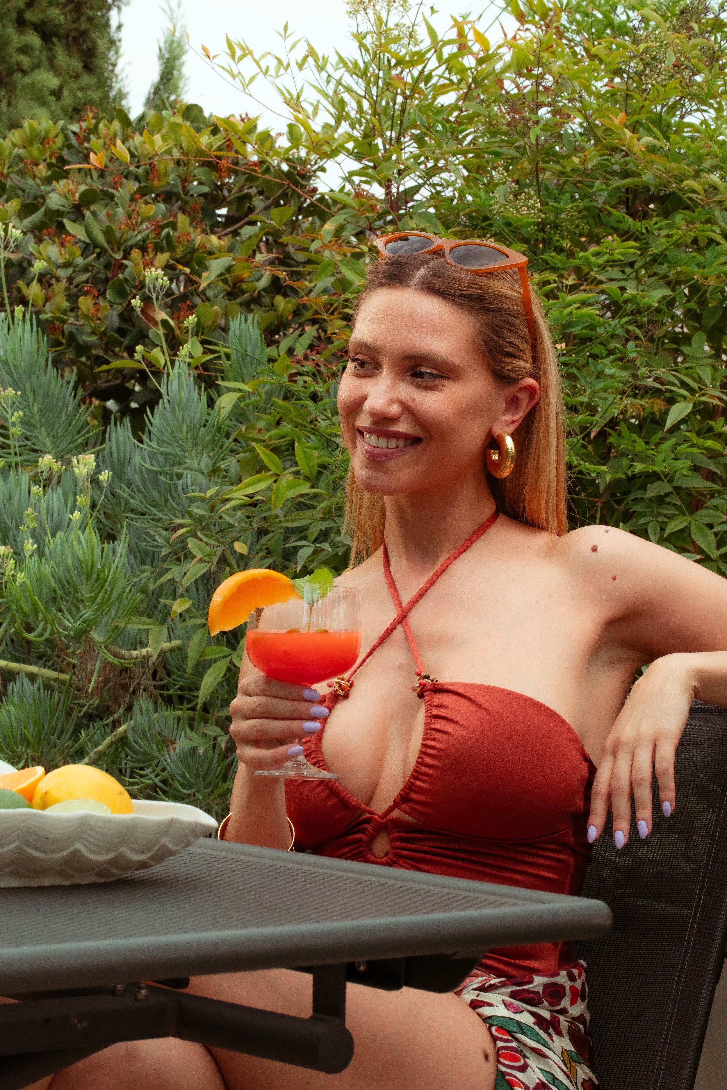Una donna sorridente beve un cocktail rosso con una fetta d'arancia, seduta in giardino con piante verdi.