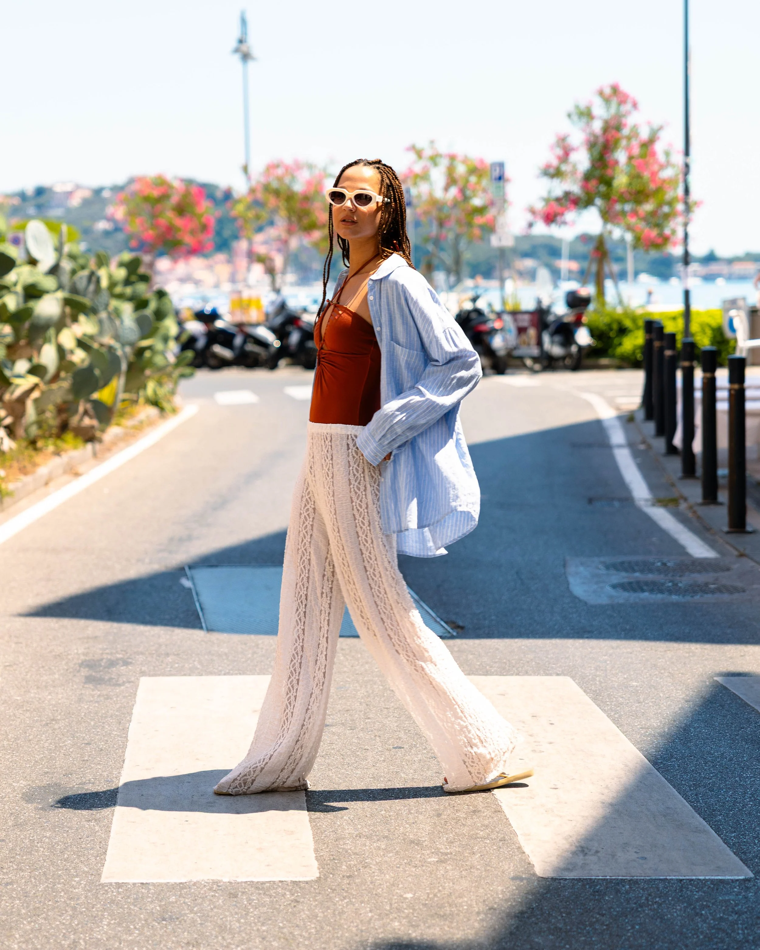 Una donna con occhiali da sole chiari, vestito marrone, pantaloni in pizzo beige e camicia a righe blu chiara, attraversa una stradapedonale vicino al mare, con alberi fioriti e barche sullo sfondo.