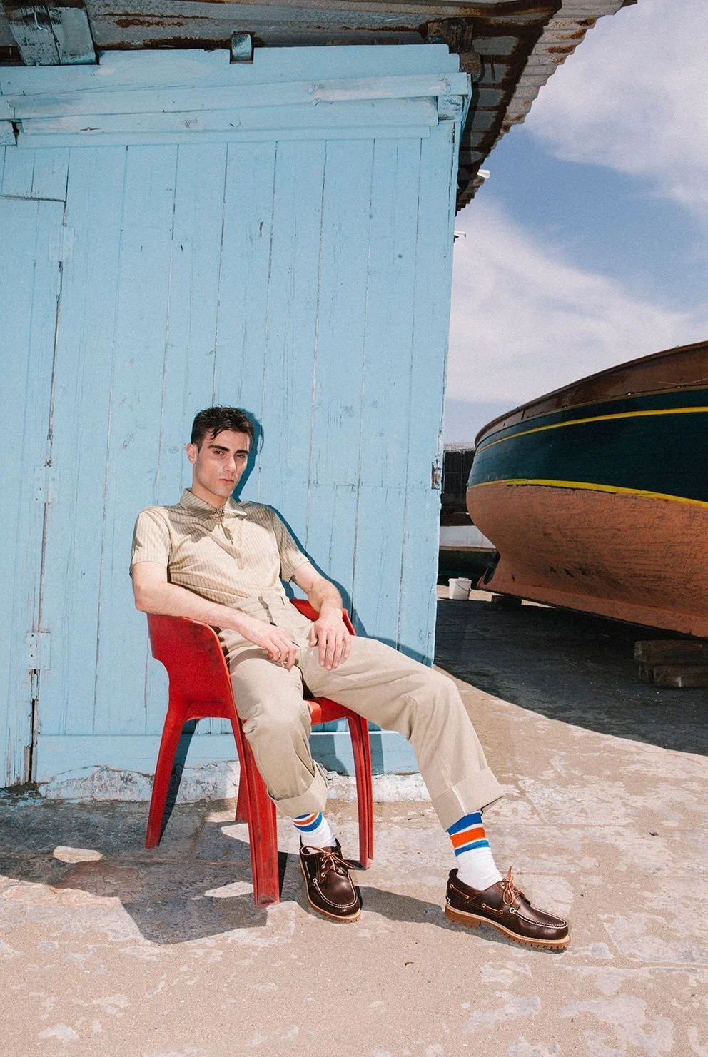 Uomo seduto su una sedia rossa davanti a un edificio di legno color azzurro, con una barca dietro di lui, sotto un cielo nuvoloso.