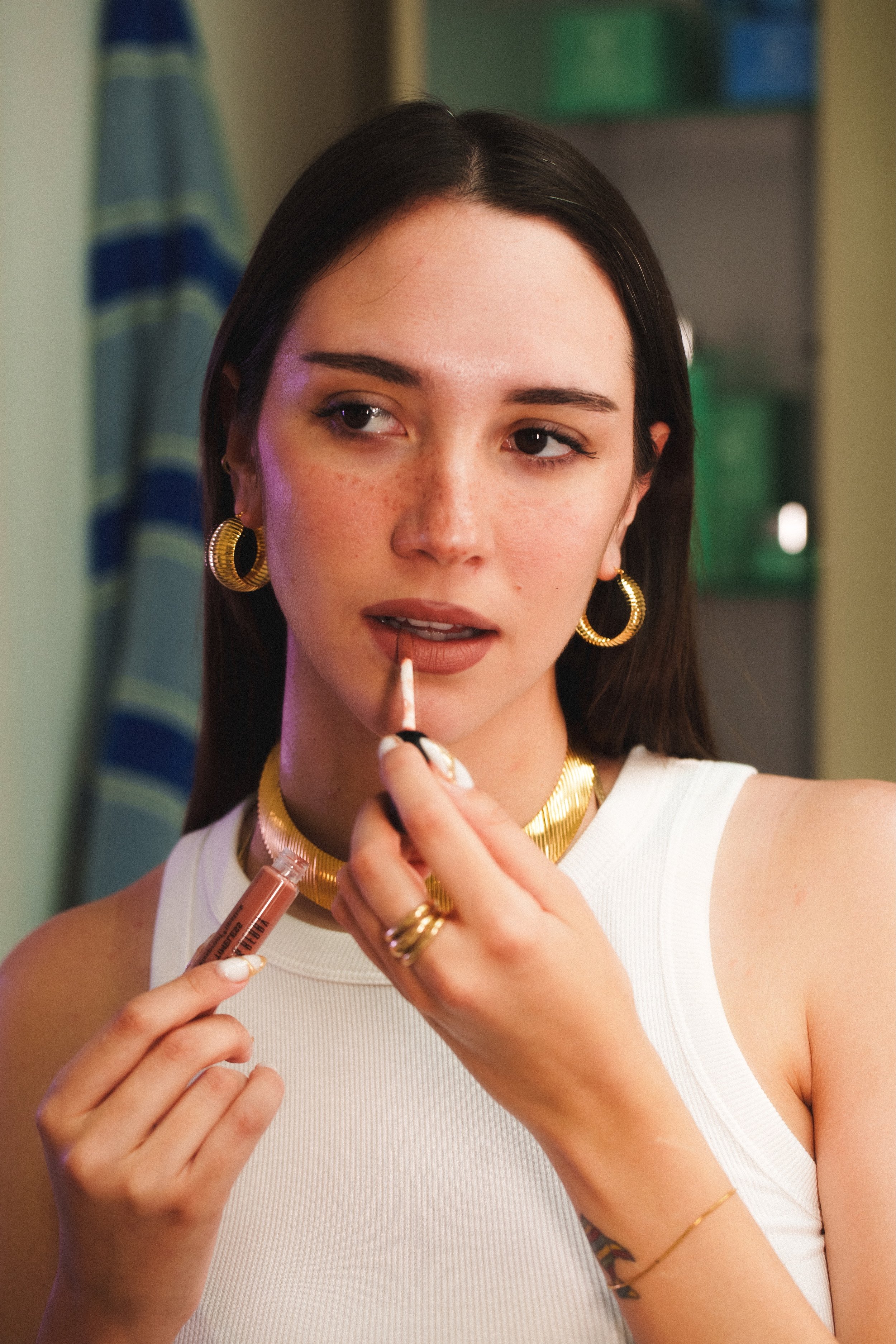 Una giovane donna con orecchini dorati e una collana sta mettendo rossetto mentre si trucca davanti a uno specchio.