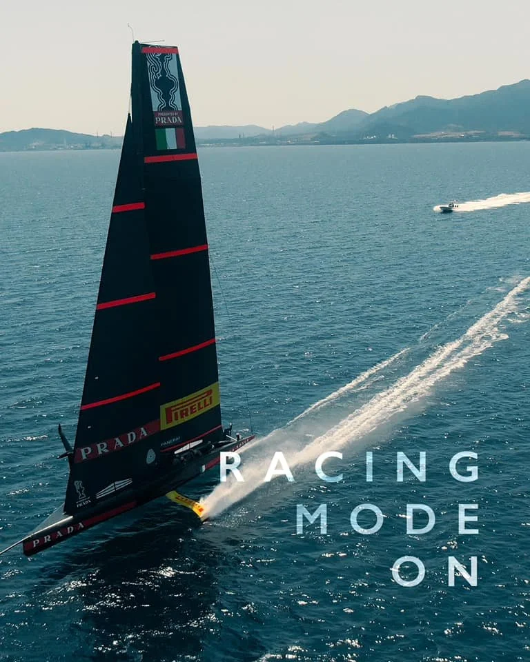 Imbarcazione da regata a vela con sponsor Prada, che naviga ad alta velocità su un mare calmo con uno sfondo di montagne, con il testo 'RACING MODE ON.'