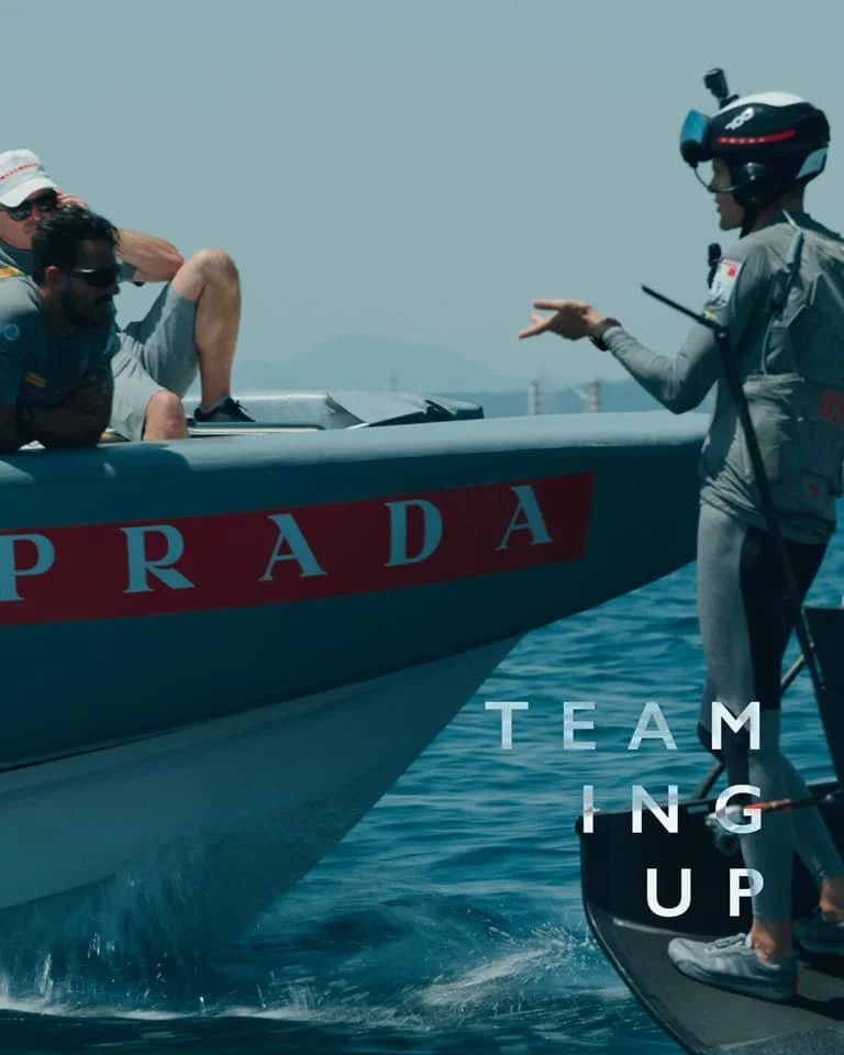 Membri del team di Prada che si preparano per una gara di barche a vela, con uno di loro che spiega qualcosa mentre gli altri ascoltano.