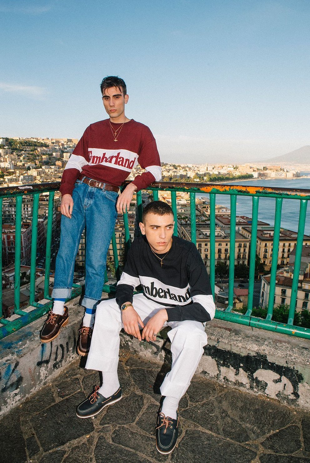 Due giovani uomini sono su una terrazza con vista sulla città e sul mare, indossano abbigliamento casual con magliette che hanno scritto 'Timberland'.