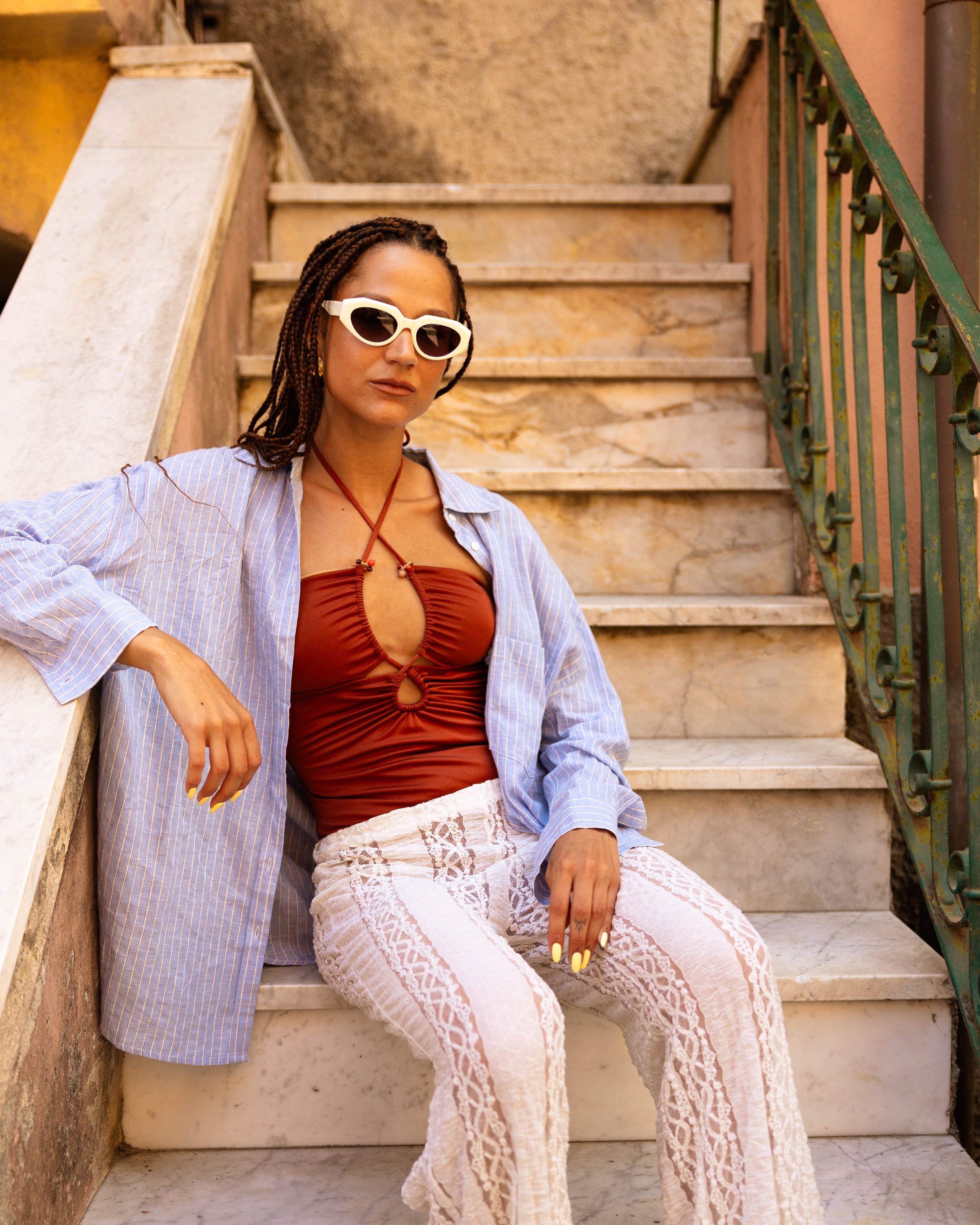 Una donna con treccine, occhiali da sole bianco, maglietta rossa, camicia azzurra e pantaloni di pizzo bianco seduta su una scala di marmo all'aperto.