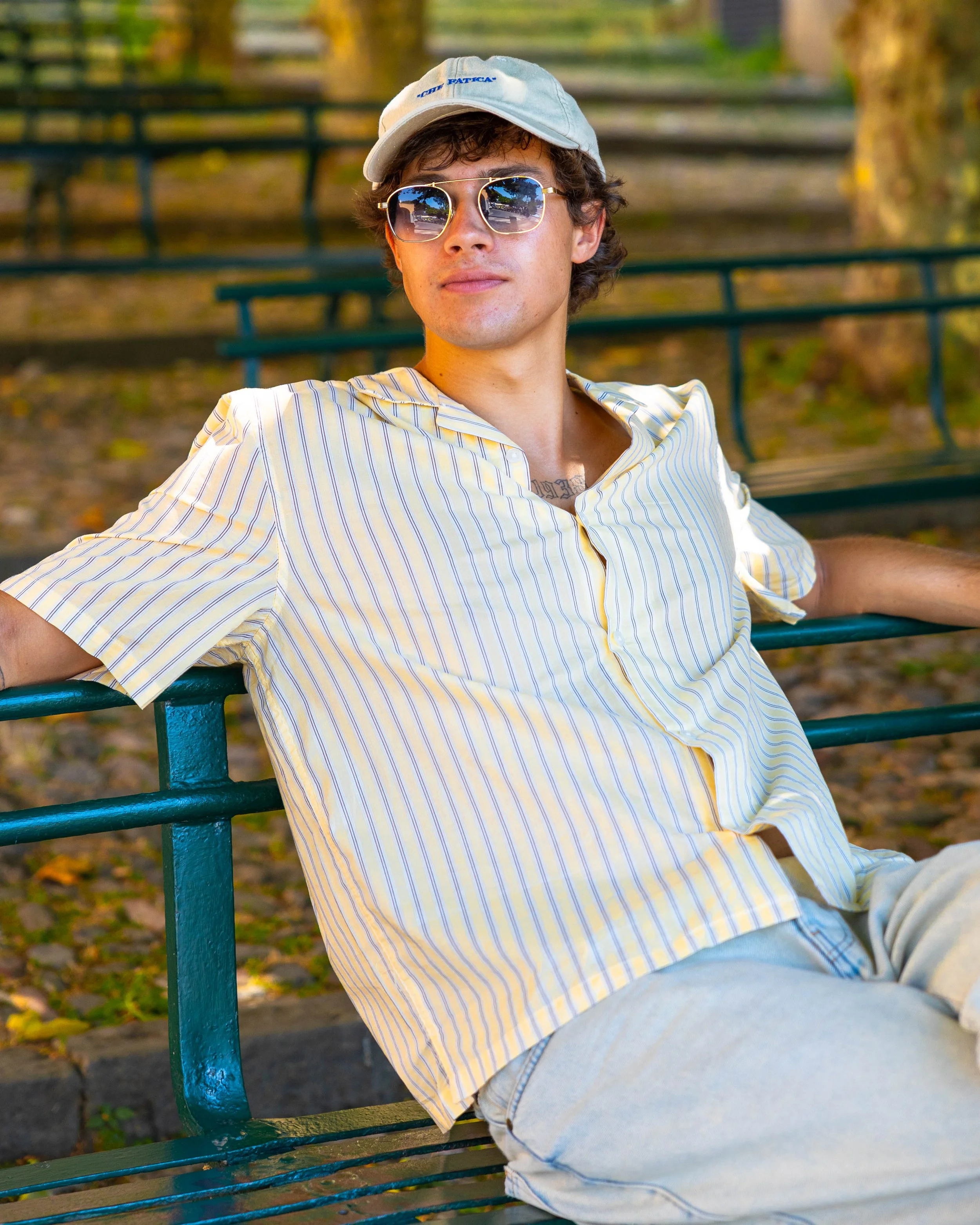 Ragazzo seduto su una panchina di ferro in un parco, indossa un cappello, occhiali da sole e una camicia a righe.