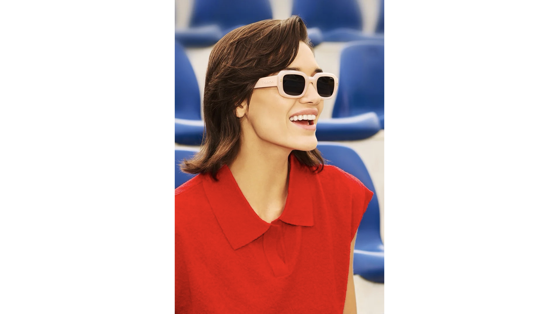 Una donna sorridente con occhiali da sole chiari e abbigliamento rosso in un stadio con sedie blu sullo sfondo.