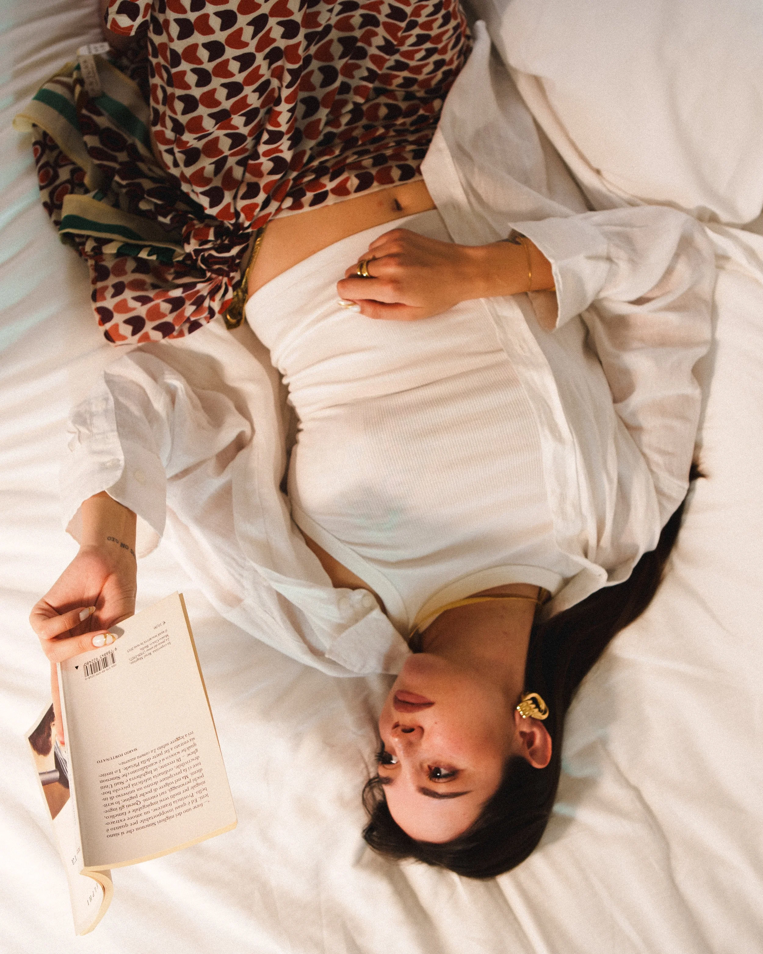 Una donna distesa su un letto, che tiene in mano un libro aperto, vestita con una maglietta bianca e una camicia di pizzo e pantaloni beige con una schiena scoperta, con orecchini dorati e anelli, in un ambiente intimo e rilassato.