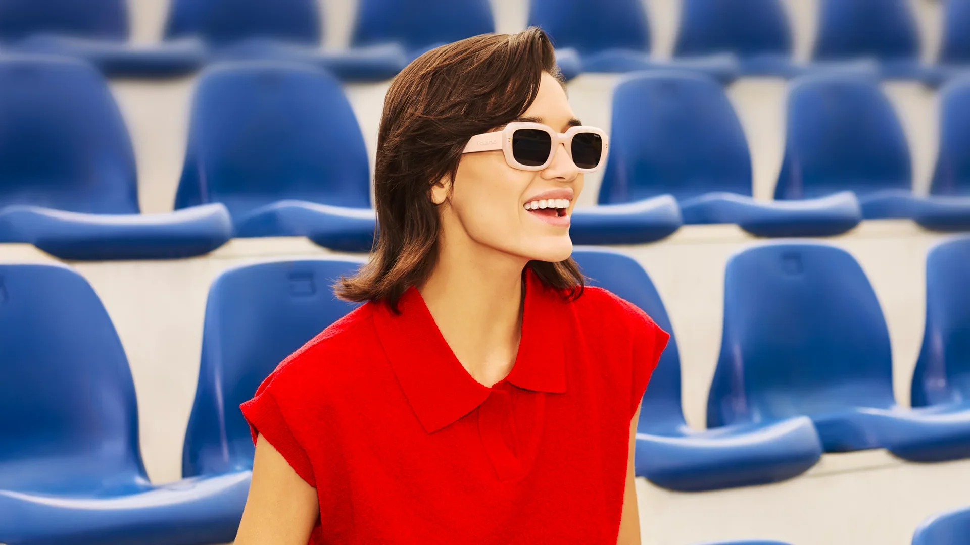 Donna con capelli castani e occhiali da sole bianchi sorride seduta in uno stadio con sedili blu.