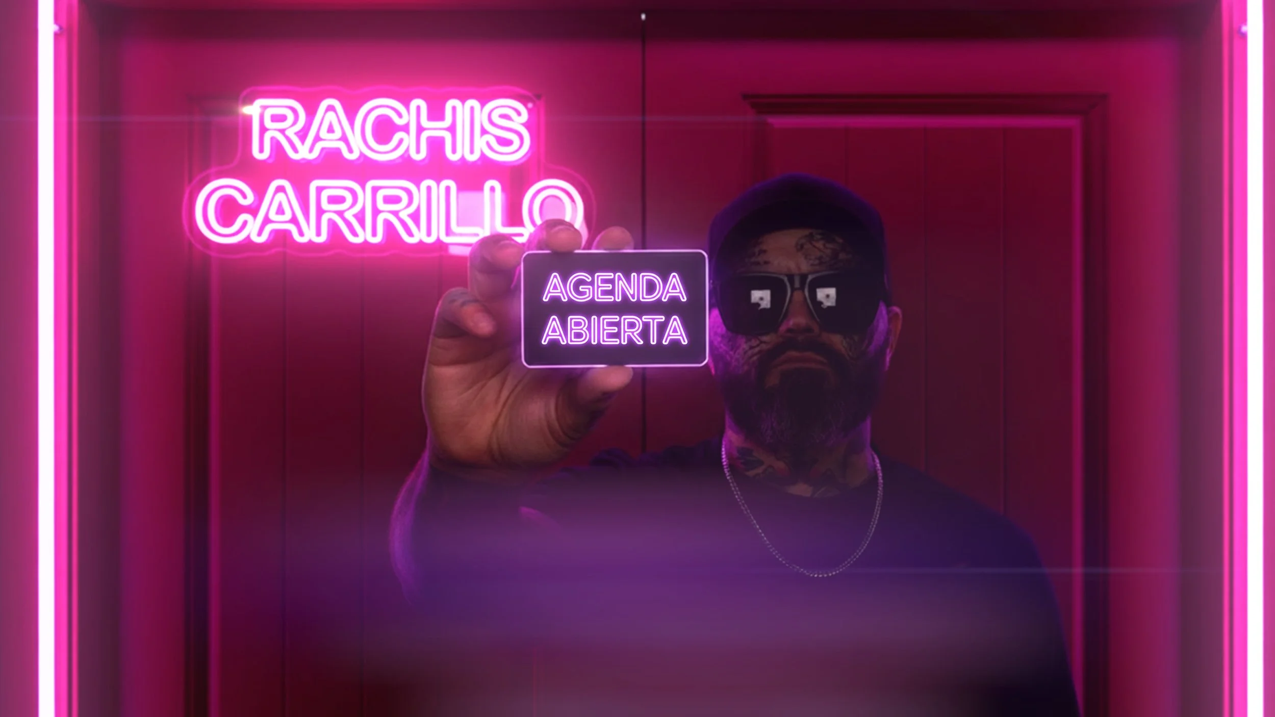 A man with tattoos, wearing a black shirt and sunglasses, holds up a small sign that reads "AGENDA ABIERTA" in front of a pink illuminated sign that says "RACHIS CARRILO." The background is a wooden paneled wall with pink neon lighting.