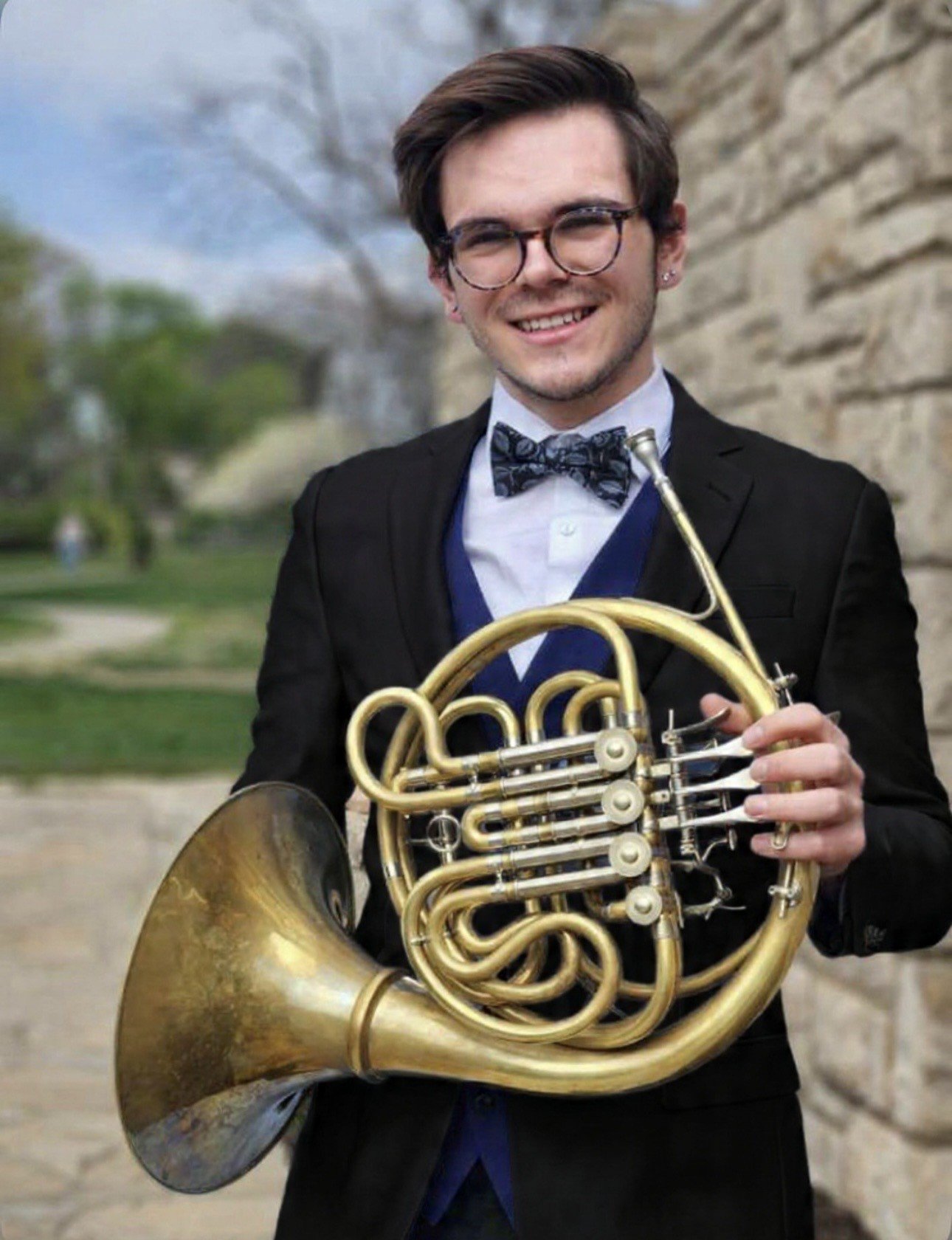 A young man wearing glasses, a black tuxedo with a bow tie, and a white shirt, smiling and holding a brass French horn outdoors near a stone wall.