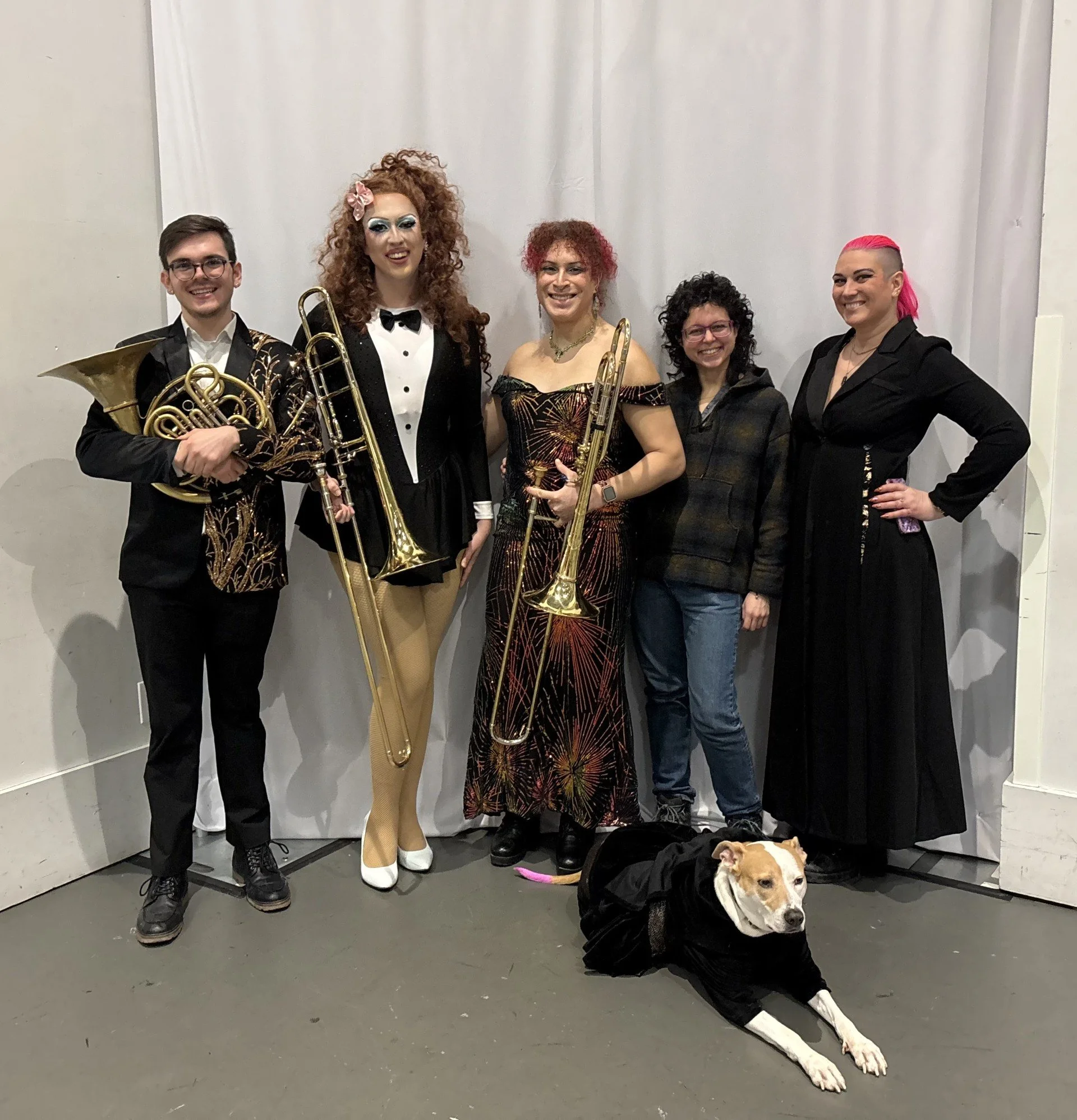 Group of five people standing together, holding musical instruments, with a dog lying on the floor in front of them. The group appears to be musicians, dressed up for a performance, and the background is a plain white curtain.