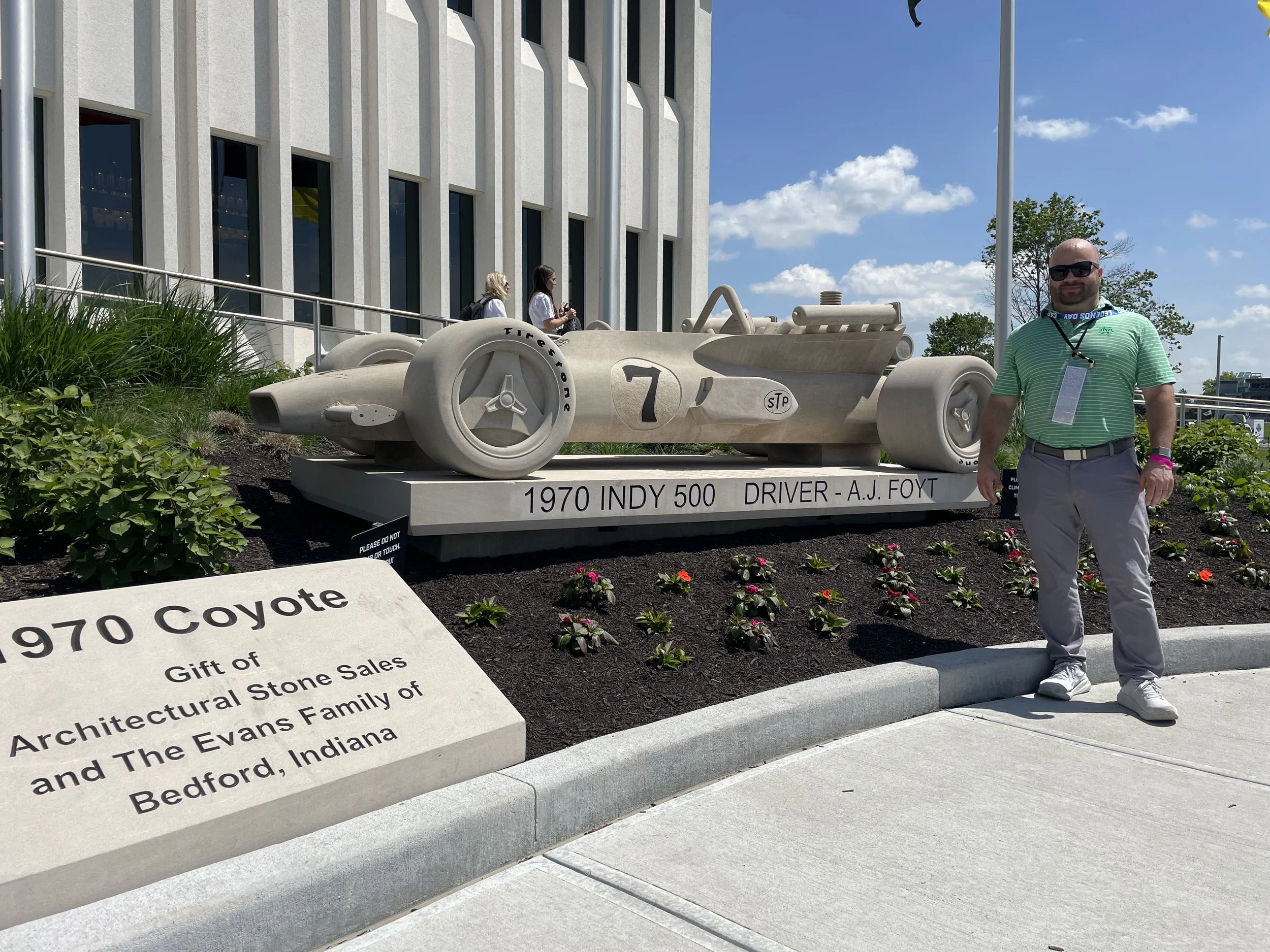 Man standing outside next to a large replica of a 1970 Indy 500 race car, which is mounted on a display platform with a sign that reads '1970 Indy 500 Driver - A.J. Foyt'. The background features a modern building, a blue sky with some clouds, and a few people walking. There is also an informational plaque in the foreground about the gift from the Evans Family of Bedford, Indiana.