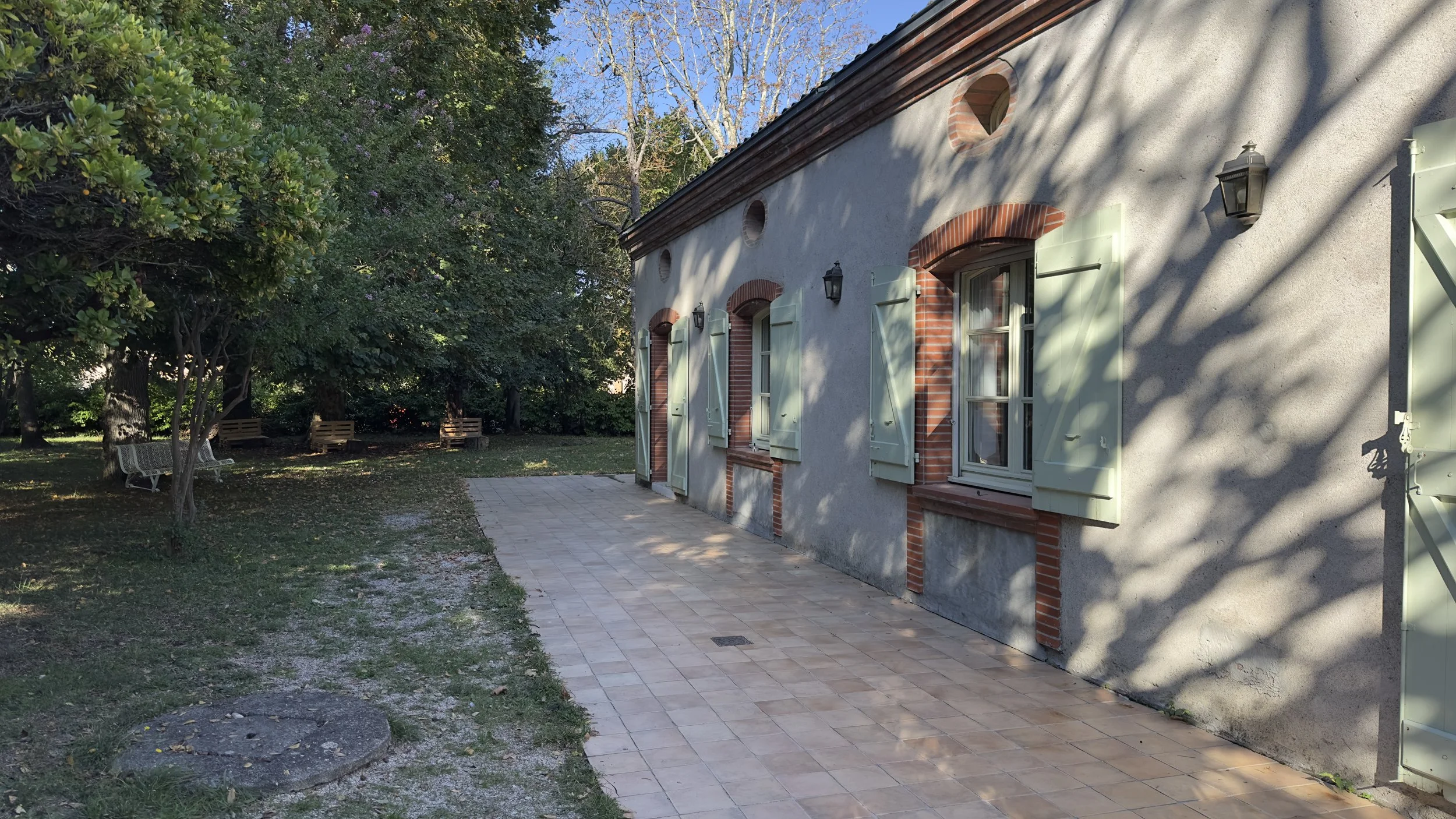 Maison avec volets verts et mur en briques, jardin avec arbres et bancs, démarche pavée."