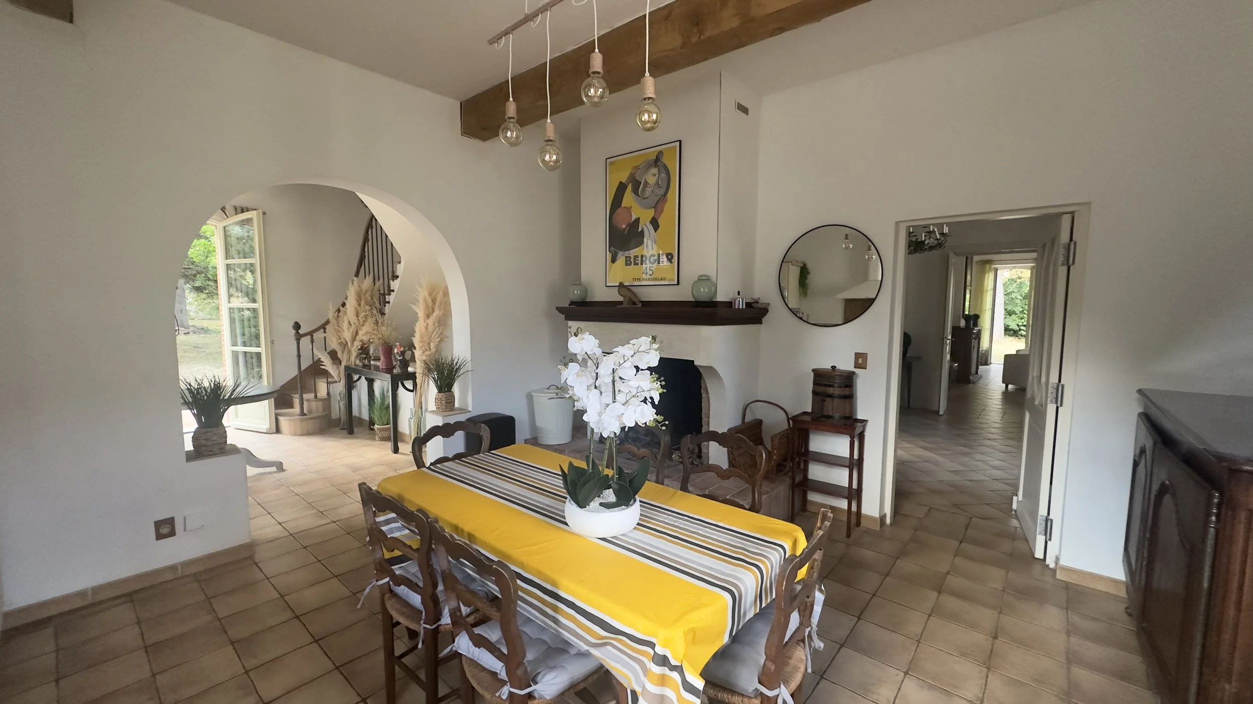 Salle à manger avec un grand tableau jaune dans la cheminée, table recouverte d'une nappe à rayures jaunes, blanches et grises, avec un vase blanc d'orchidées blanches. Ouvertures sur un jardin avec plantes et lumière naturelle.