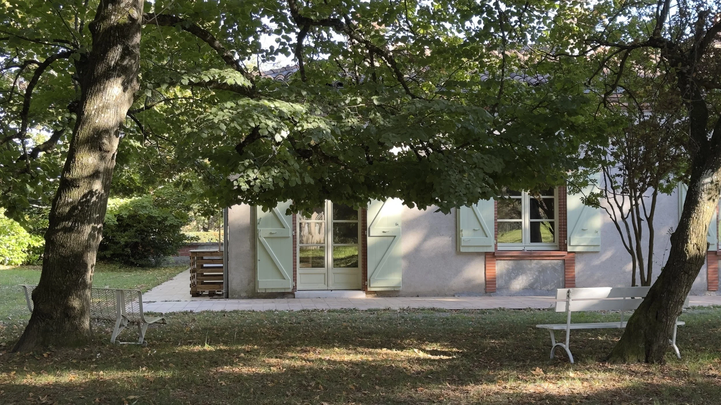 Une maison avec des fenêtres et volets verts, entourée d'arbres, avec des bancs dans le jardin.