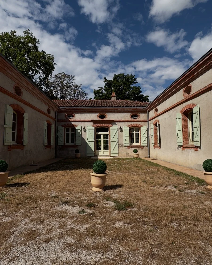 Cour intérieure d'une maison en pierre avec volets verts et pots de buis, ciel partiellement nuageux.