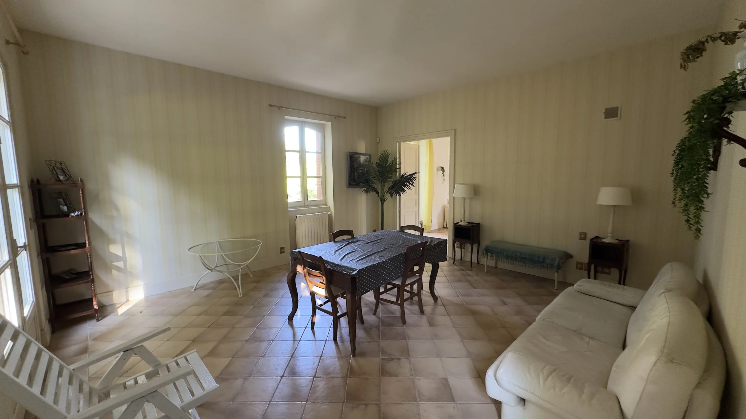 Salon avec table en bois, canapé beige, étagère en bois, lampes, et décoration végétale, lumineux par fenêtres.