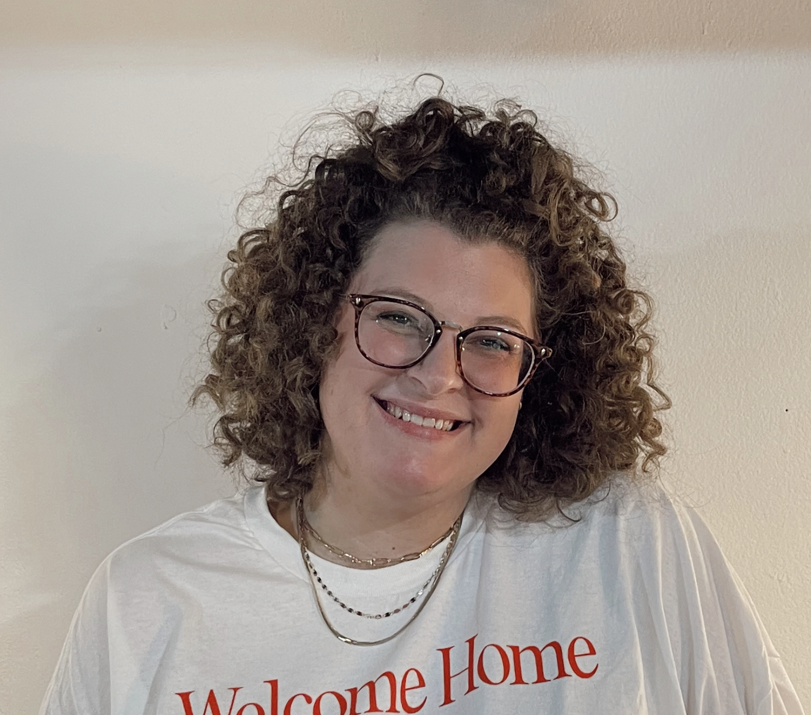 Close-up of a smiling woman with curly hair, wearing tortoise shell glasses and a white blouse, standing in front of a brick wall.