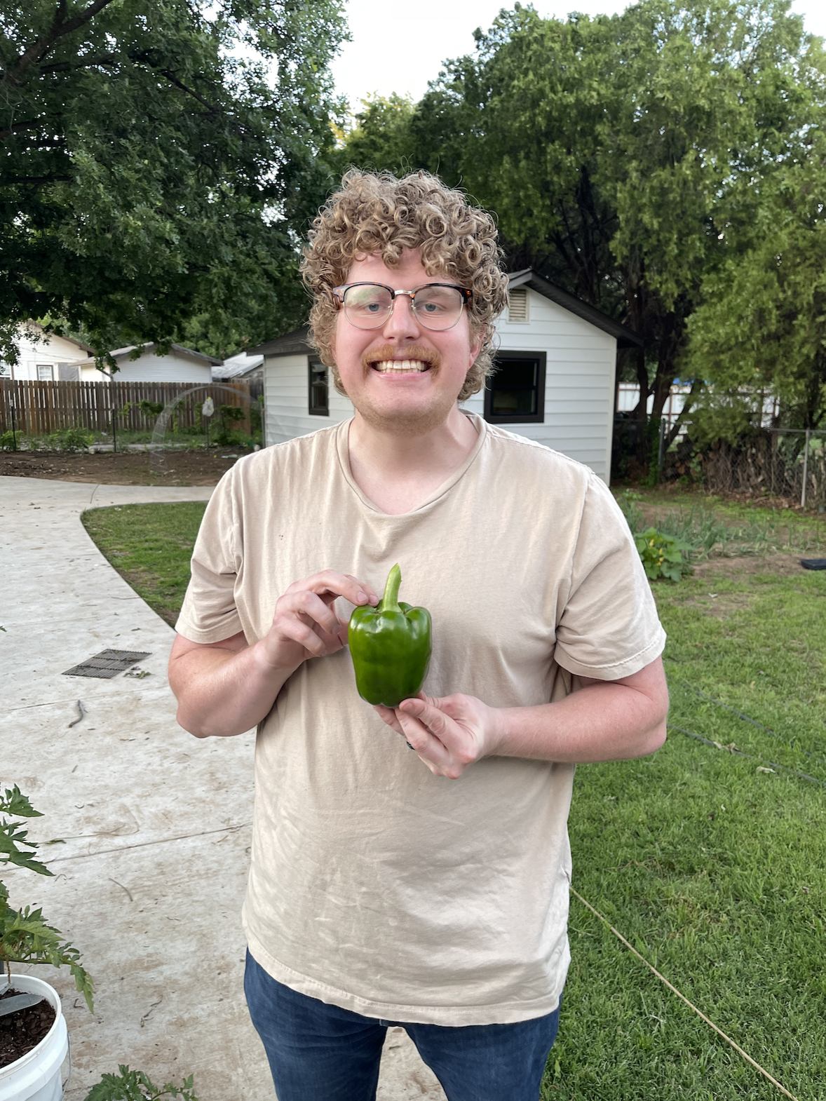 Portrait of a smiling man with curly hair and glasses, holding a green bell pepper that he grew from his garden.