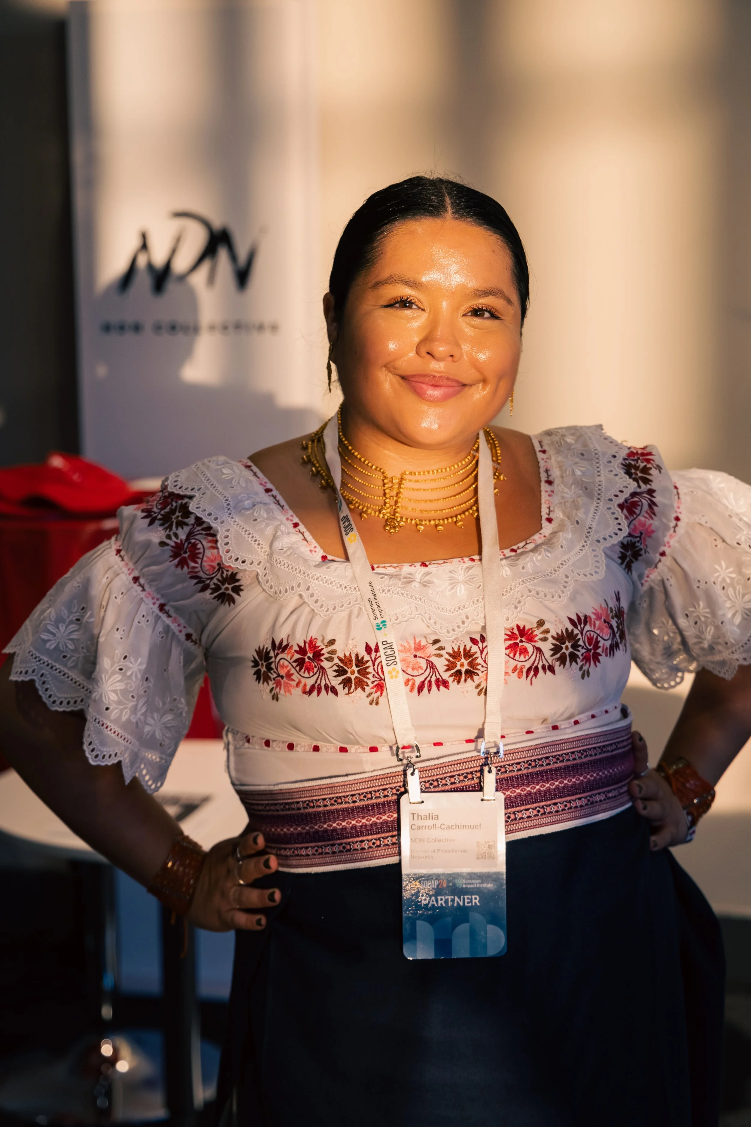 Woman smiling wearing traditional Mexican embroidered blouse and gold jewelry at a conference, with a name badge around her neck.