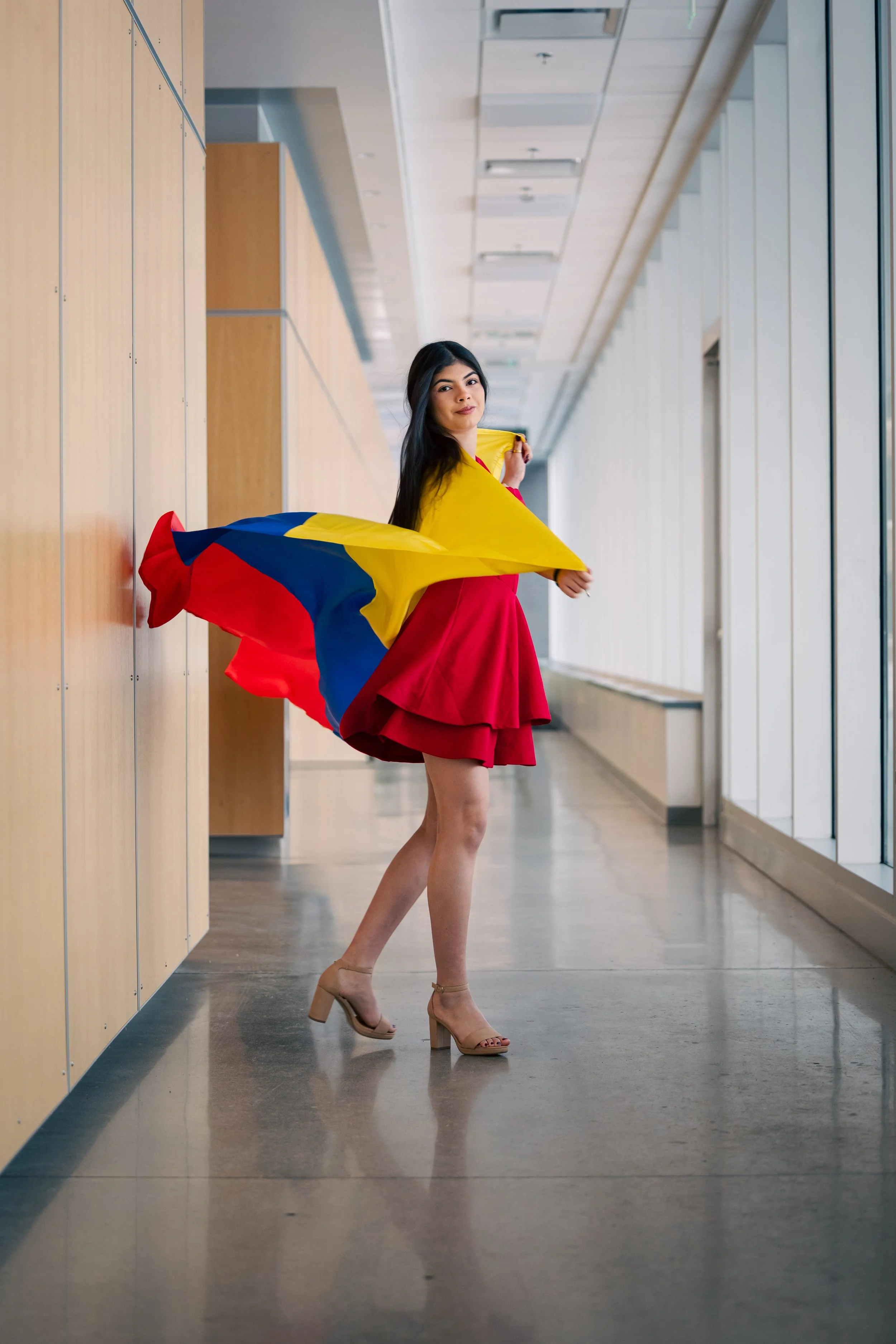 A woman is standing in a modern indoor corridor, wearing a red dress and beige high heels, holding a flag with yellow, blue, and red colors.