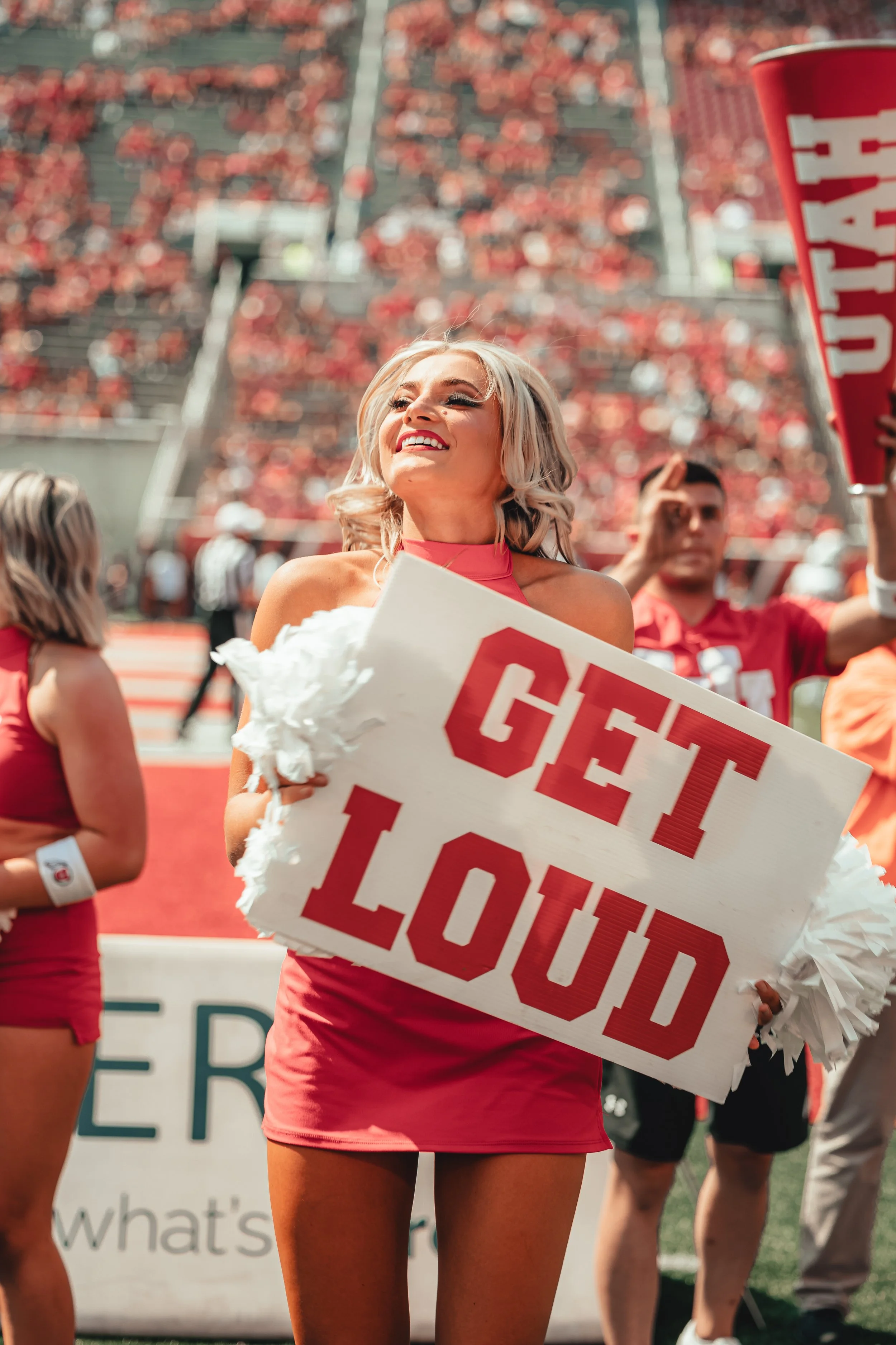 Cheerleader in a red outfit smiling and holding a sign that says "GET LOUD" during a football game.