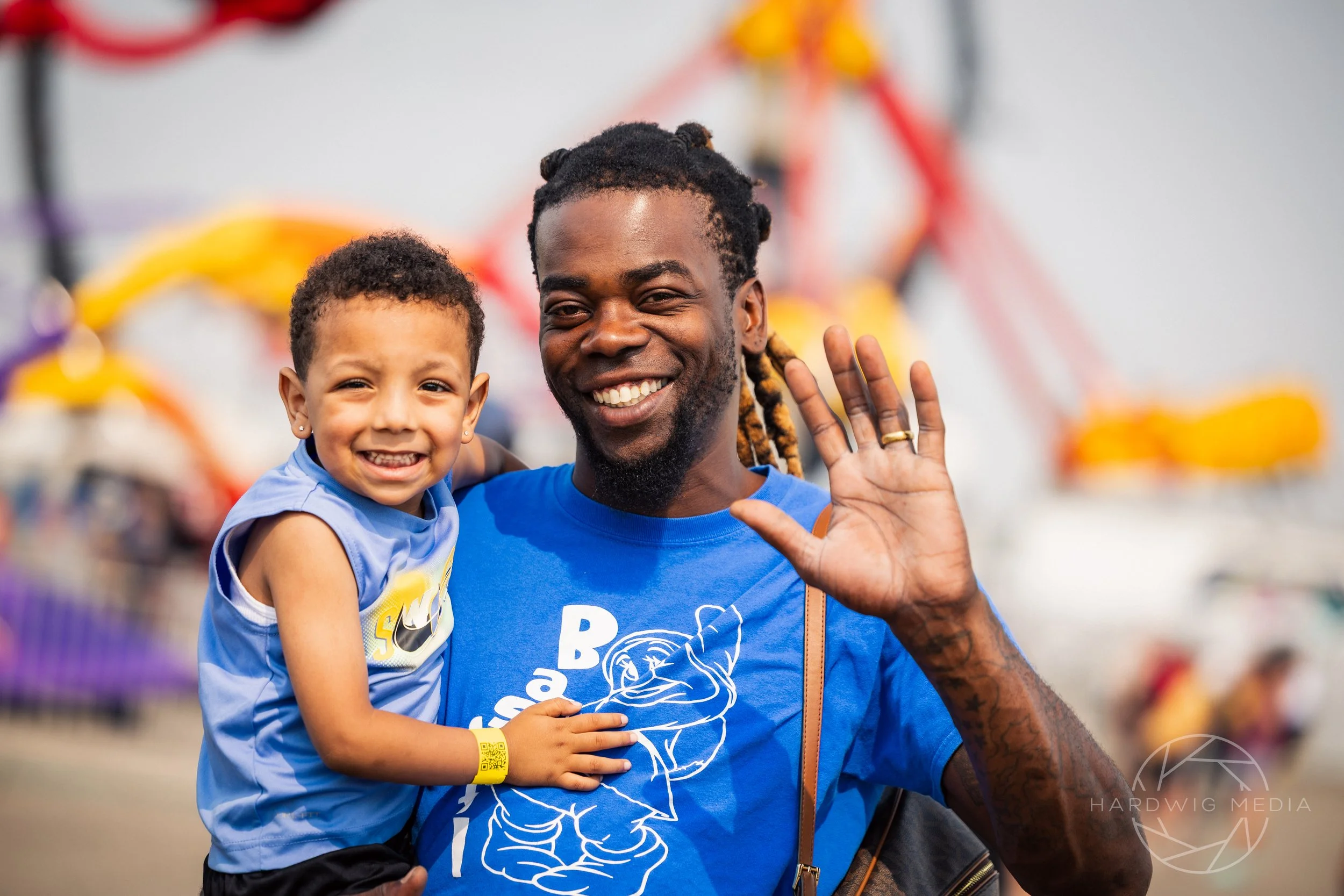 A man and a child at an amusement park, both smiling and waving, with rides in the background.