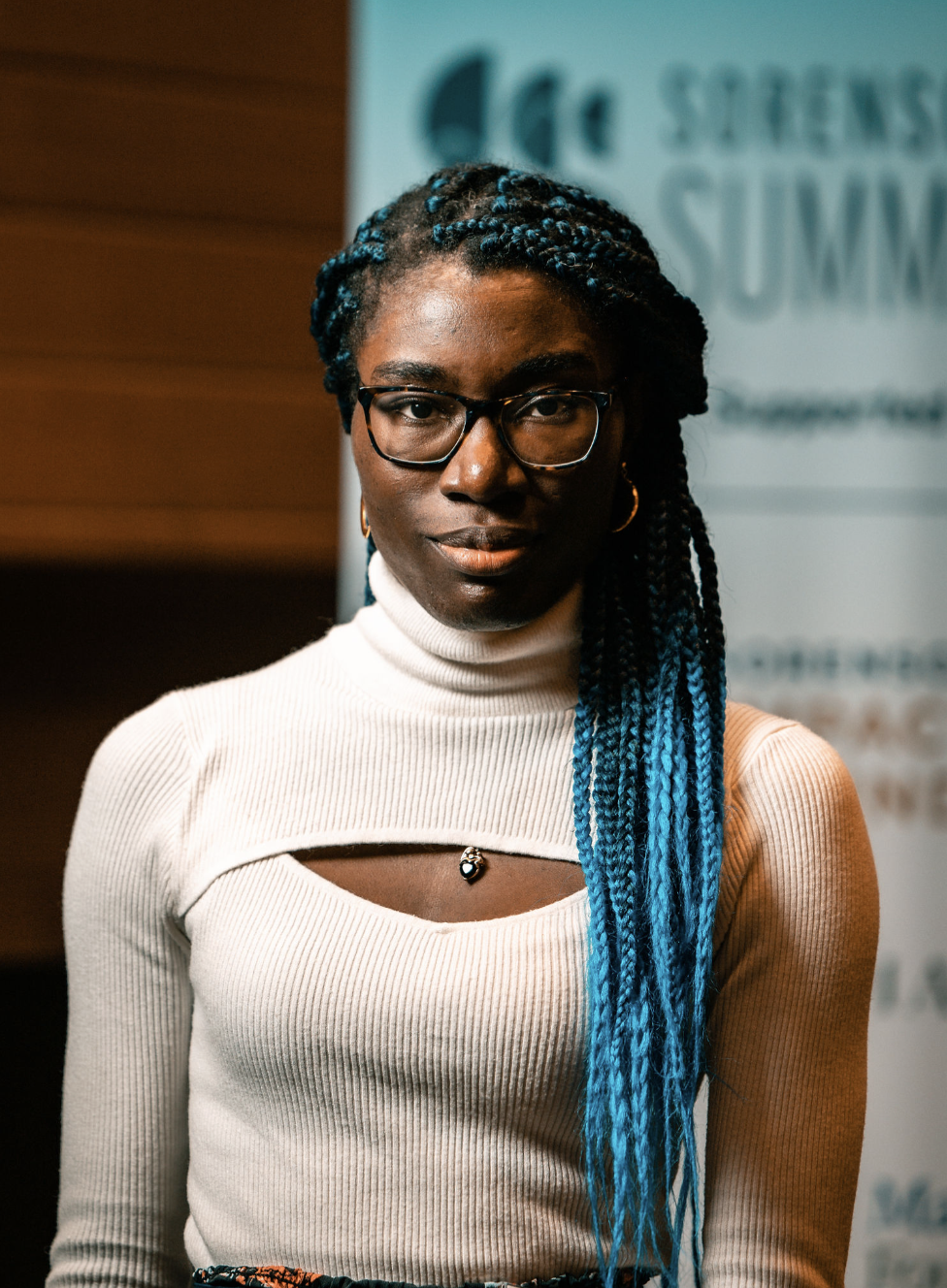 A young woman with dark skin, curly blue-tinted braids, glasses, and gold hoop earrings, wearing a cream-colored turtleneck sweater with a cut-out detail at the chest, standing indoors in front of a blurred background with a blue sign.