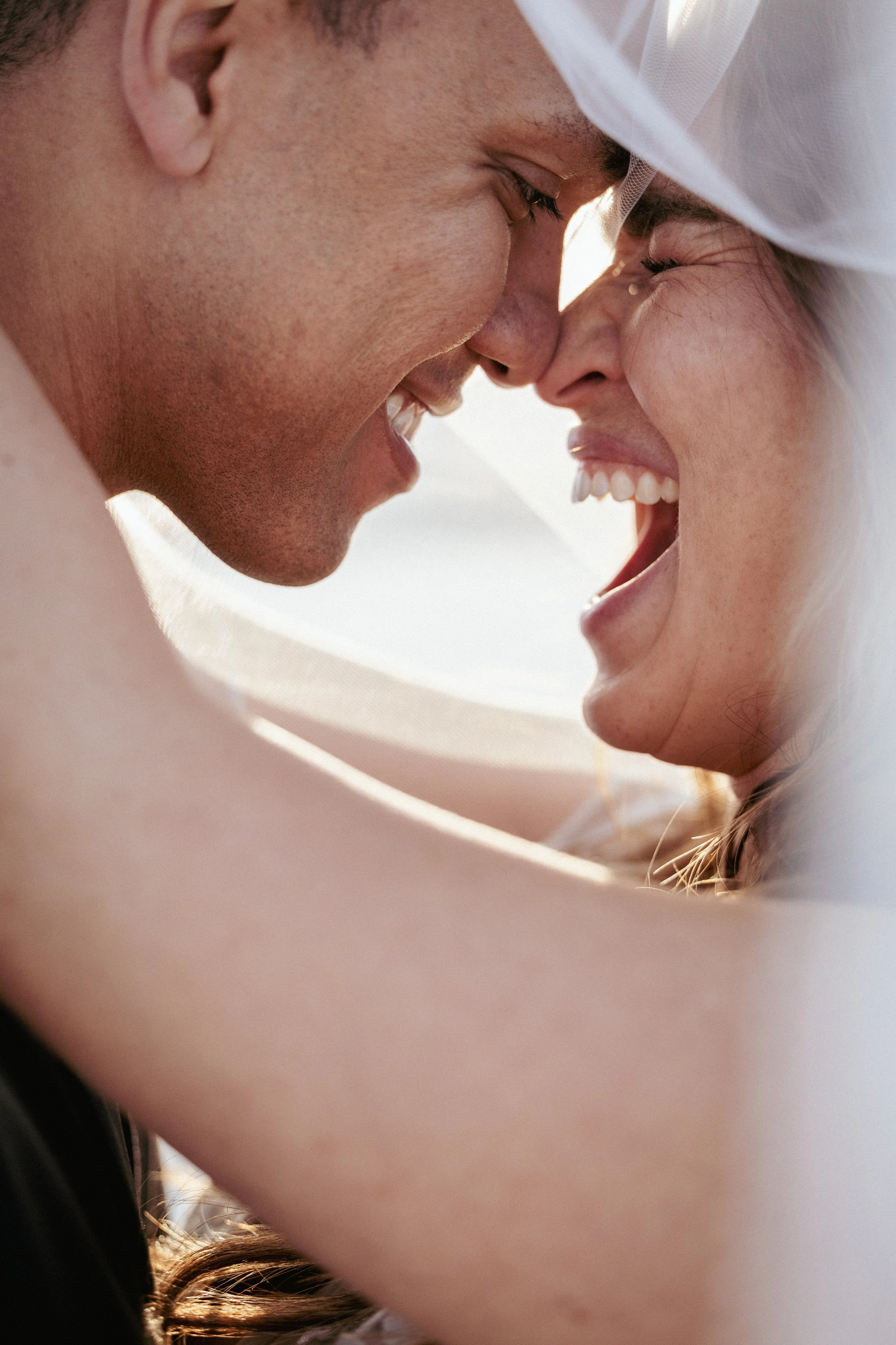 Close-up of a couple smiling and laughing with their foreheads and noses touching, partially covered by a white veil.