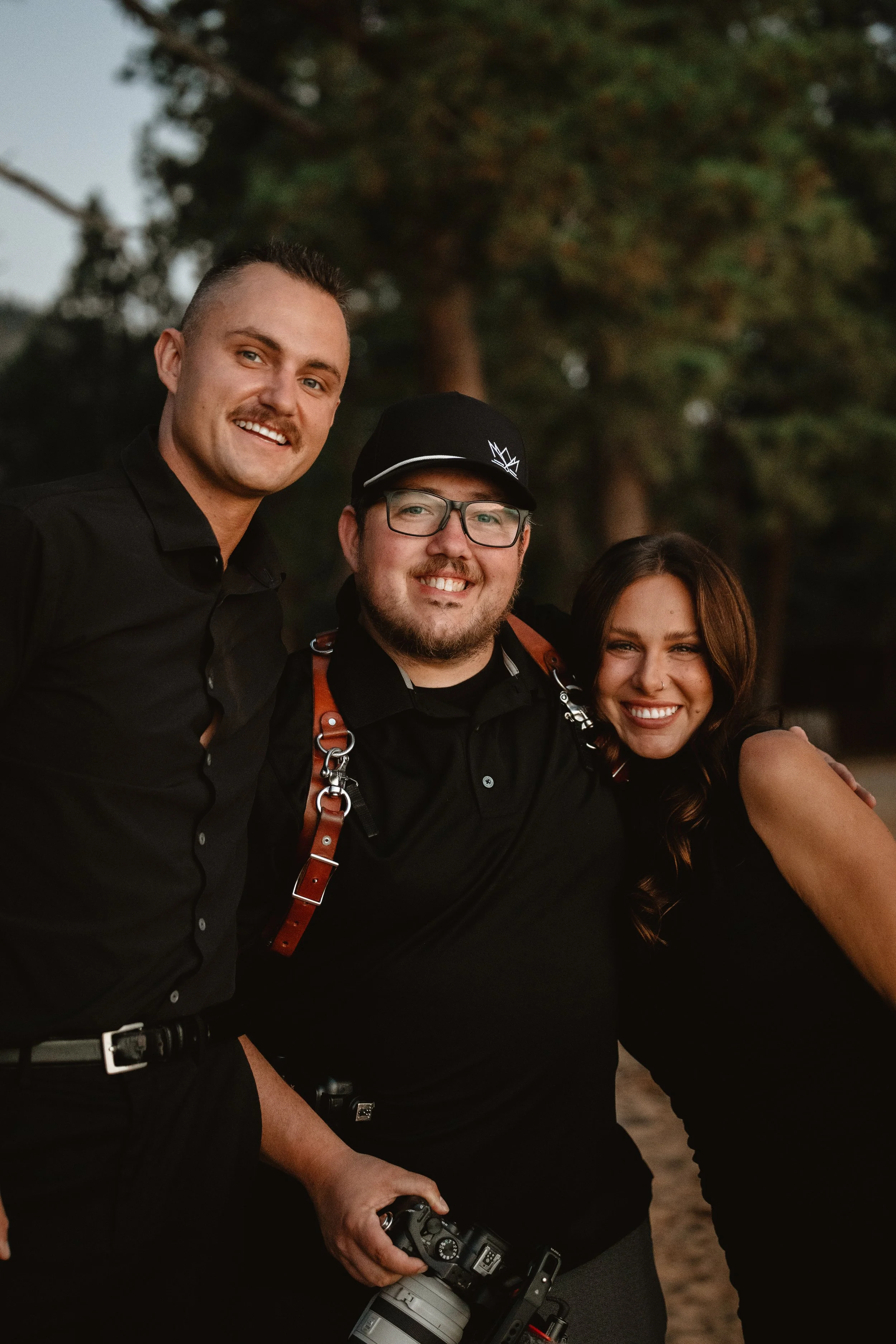 Three friends smiling and posing outdoors at sunset, with trees in the background. One person is holding a camera.