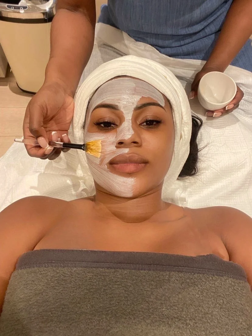 A woman receiving a facial treatment, with a skincare mask being applied on her face, lying on a spa bed, with a towel wrapped around her chest and a headband keeping her hair back.