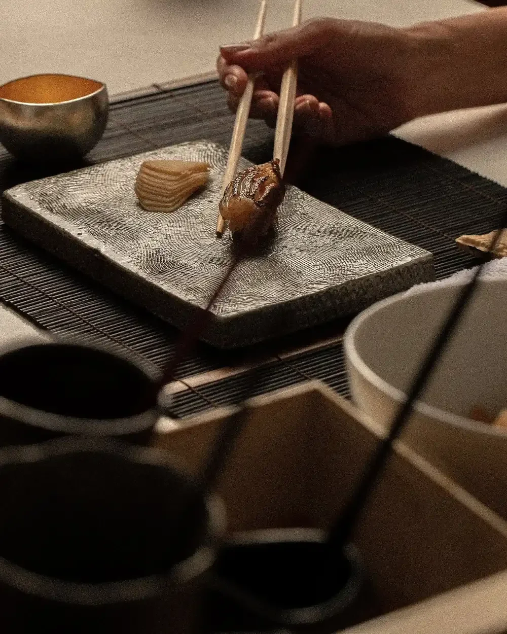 A hand lifting nigiri with chopsticks from a textured stone plate.