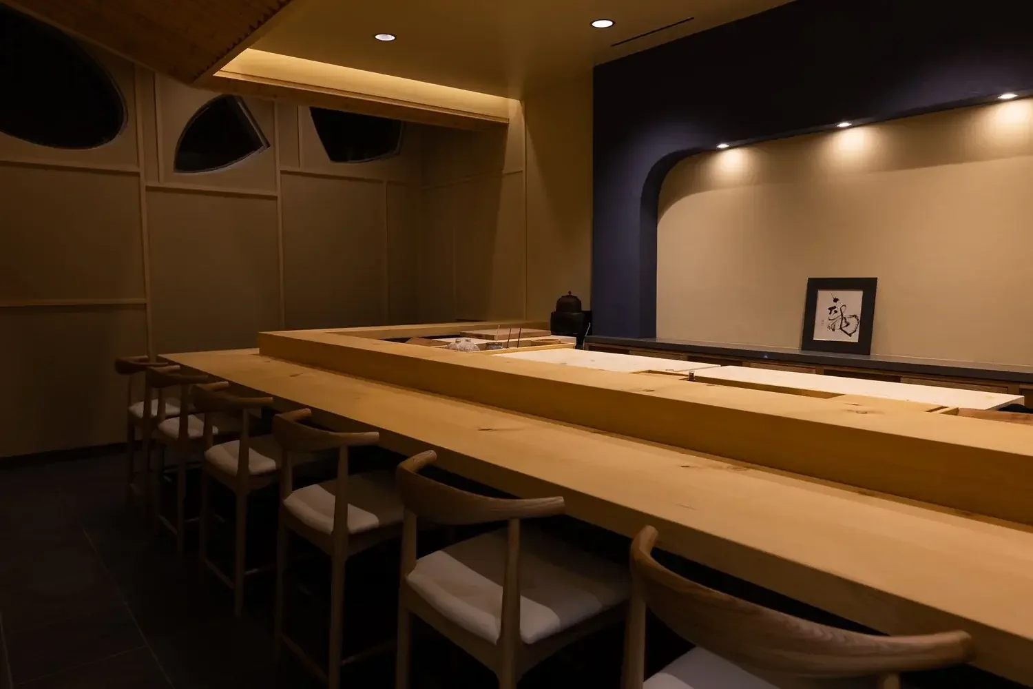 Minimalist wooden sushi counter with white chairs in a dimly lit restaurant.