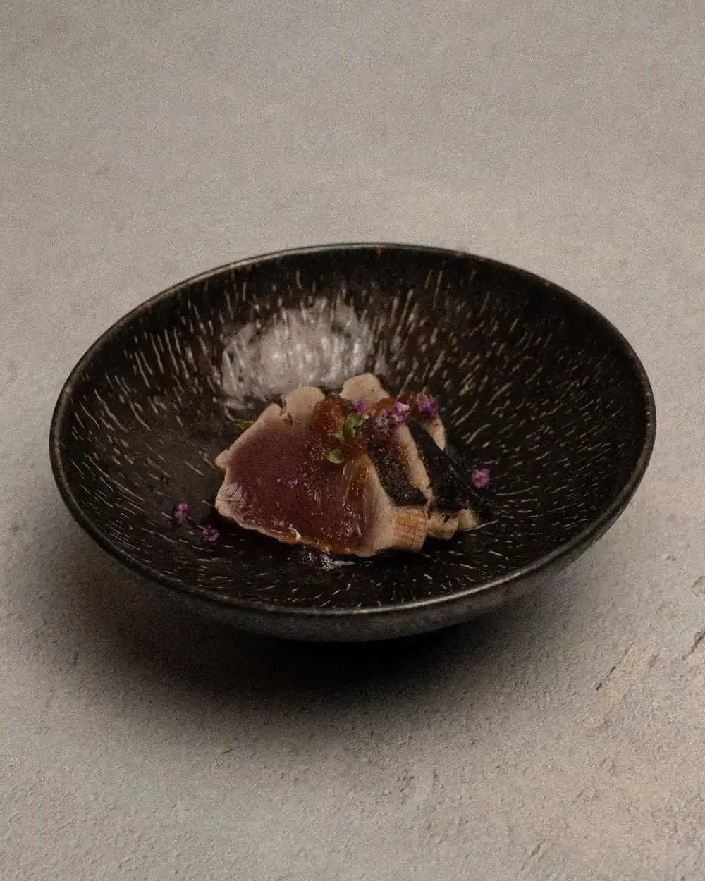 Seared Katsuo no Tataki slices garnished with purple flowers in a dark coconut bowl.