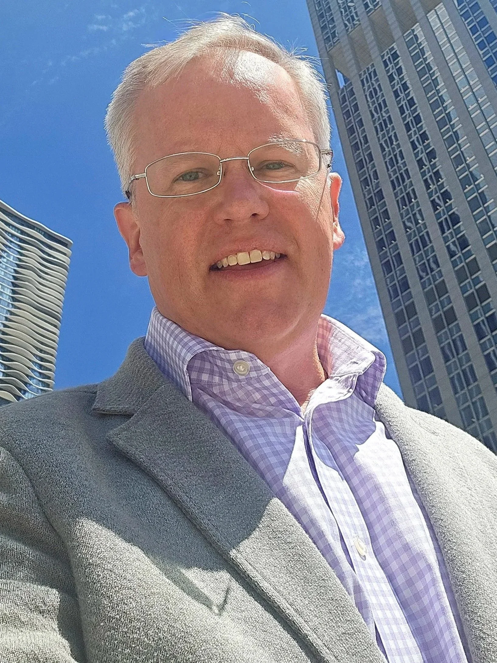 Karl Rectanus wearing glasses and a light gray blazer, smiling, in a city with tall modern buildings and a clear blue sky.