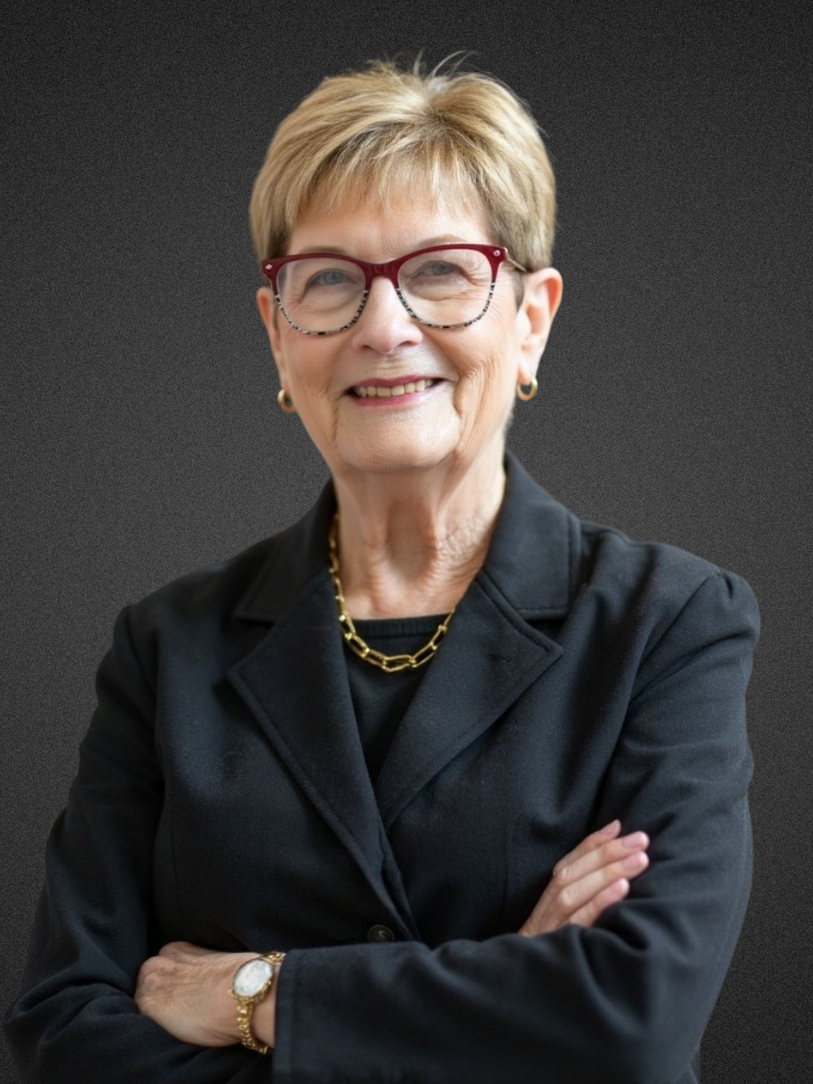 An older woman with short blonde hair, wearing red and black glasses, gold jewelry, and a black blazer, standing with arms crossed against a blue, textured background.