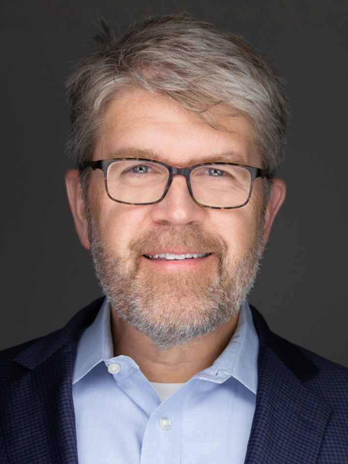 A middle-aged man with gray hair and beard wearing glasses, a light blue shirt, and a dark blazer, smiling against a dark background.