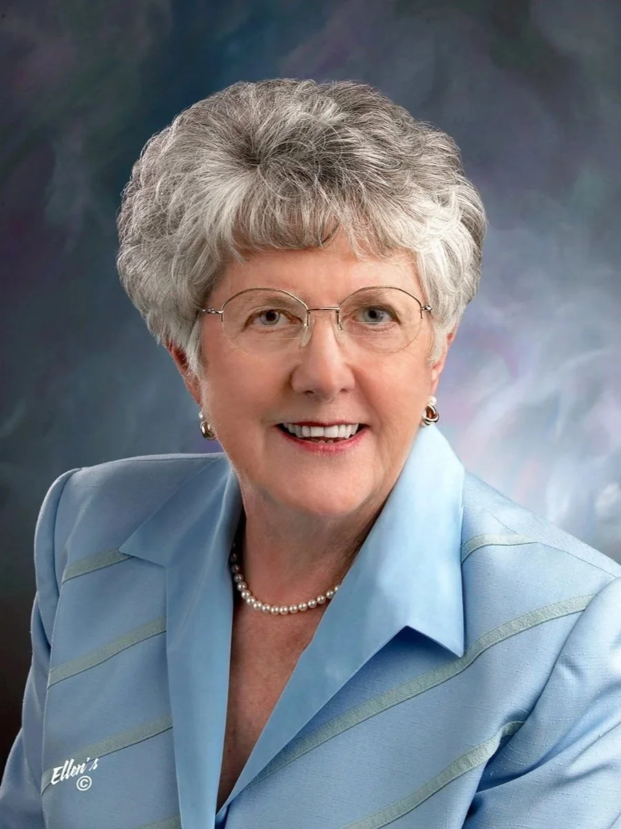 A portrait of an elderly woman with short, curly gray hair, glasses, pearl earrings, a pearl necklace, and a light blue blazer, smiling against a dark, cloudy background.