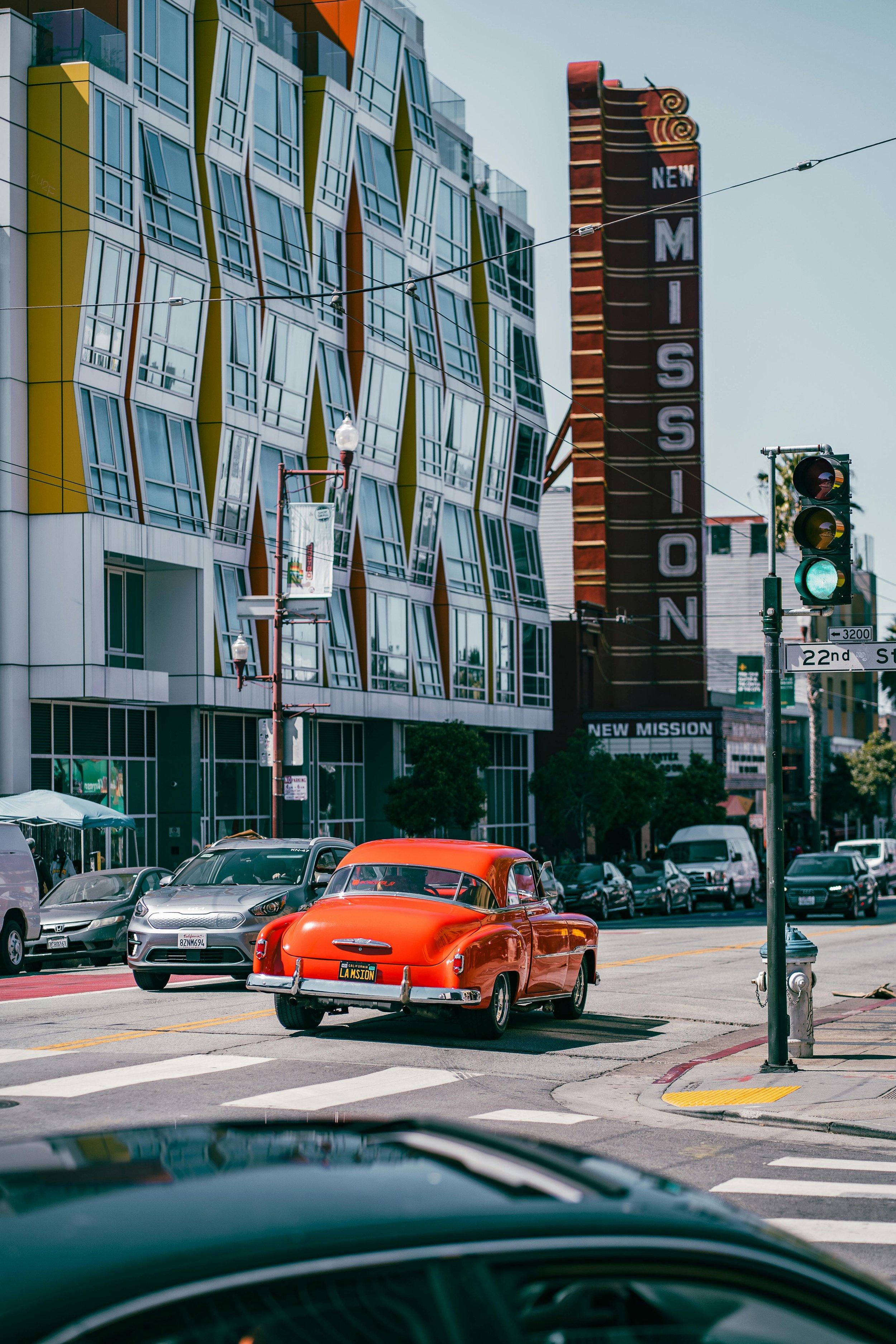 City street scene showing a vintage red car with California license plate, modern cars, a street light, and colorful buildings, including a large vertical sign that reads 'NEW MISSION'.