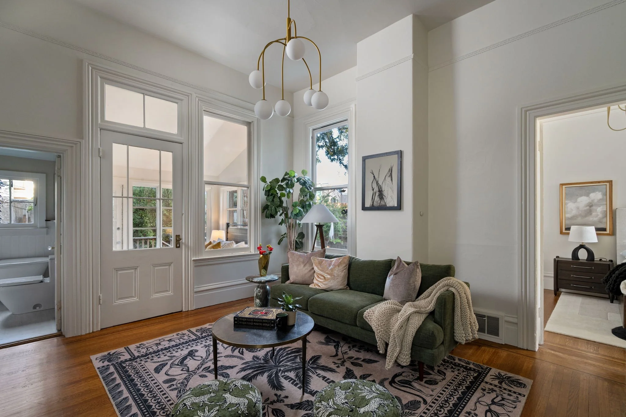 Living room with green sofa, patterned rug, coffee table, potted plant, window, and doorway to a bedroom.