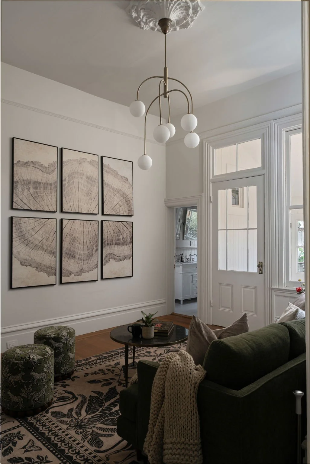 Living room with white walls, large windows, and a white door leading to a bathroom. Framed artwork of a tree cross-section hangs on the wall. A green sofa with pillows and a chunky knit blanket faces a black coffee table. Two patterned ottomans are positioned near the sofa. A modern chandelier with white globe lights hangs from the ceiling, and a patterned rug covers the floor.