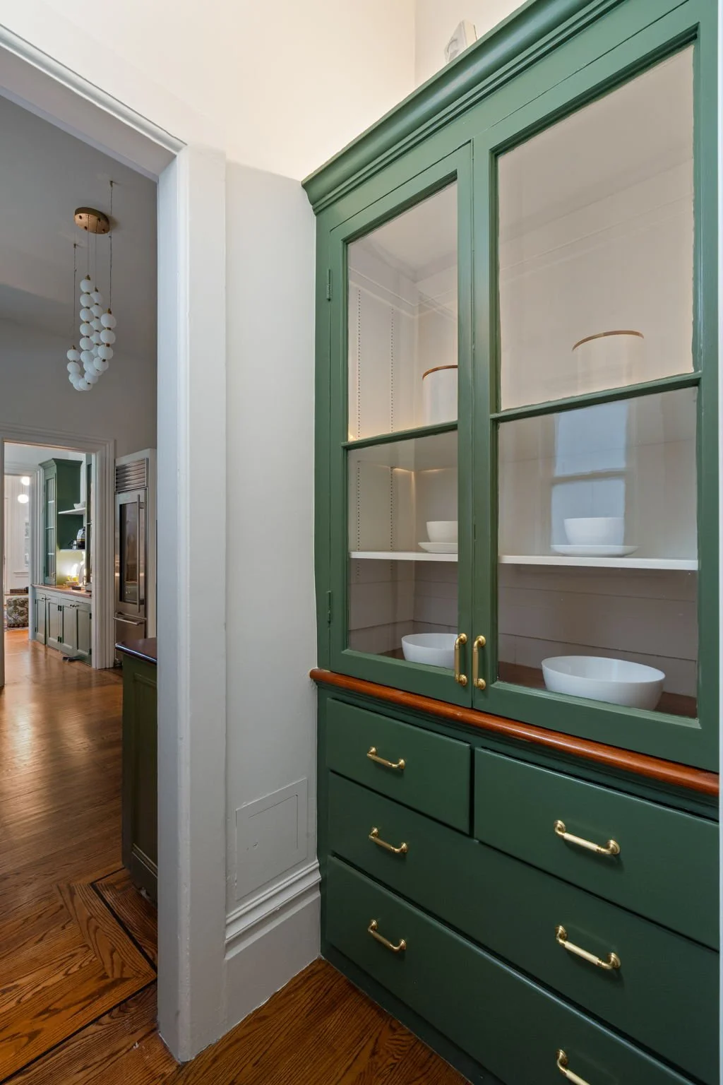A green china cabinet with glass doors, brass handles, and drawers, displaying white bowls inside, in a room with white walls and hardwood floors.