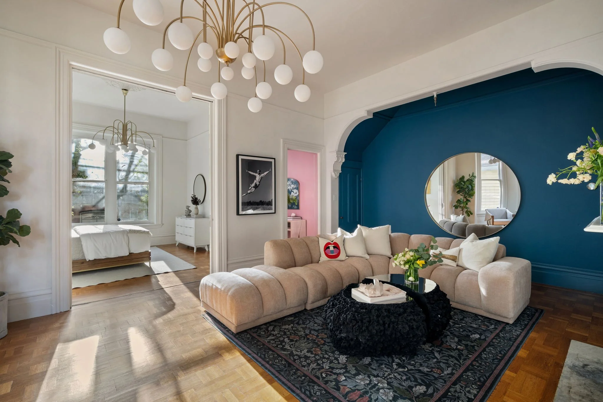 A living room with a blush pink curved sofa, black textured throw blanket, and a black coffee table with a floral arrangement and decor. The space features a bright blue accent wall with a round mirror, white walls, and hardwood floors. There's a lar