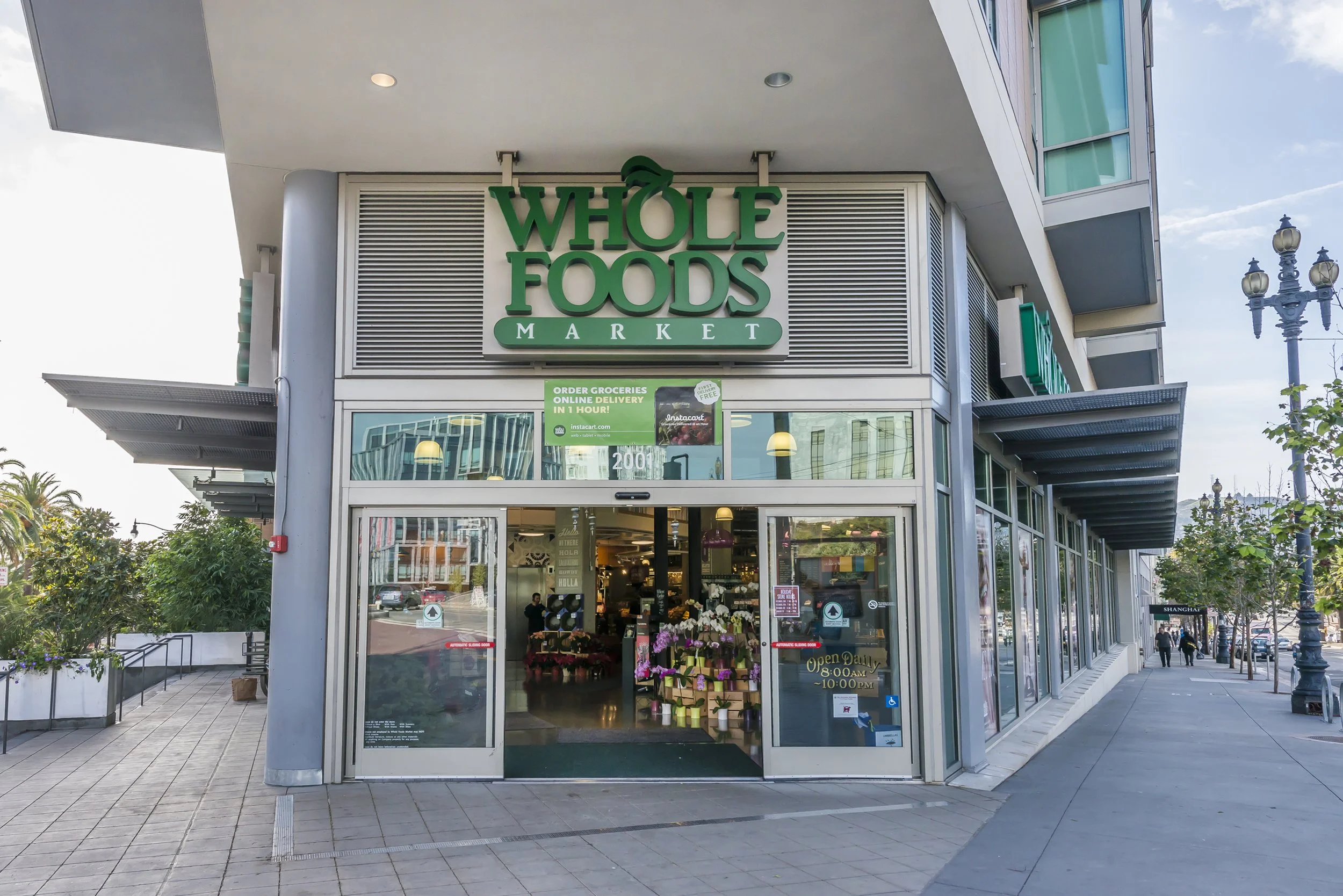 Exterior view of a Whole Foods Market store with large green sign and glass entrance, flowers displayed outside, and a sidewalk in front.