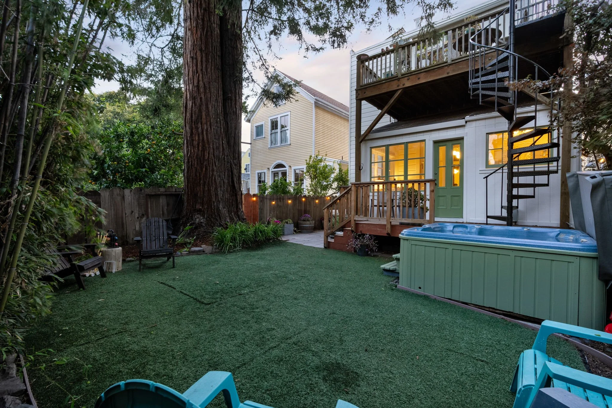 Backyard with outdoor seating, a large tree, a fenced garden, a play tub, and a house with lit windows and a deck with stairs at dusk.