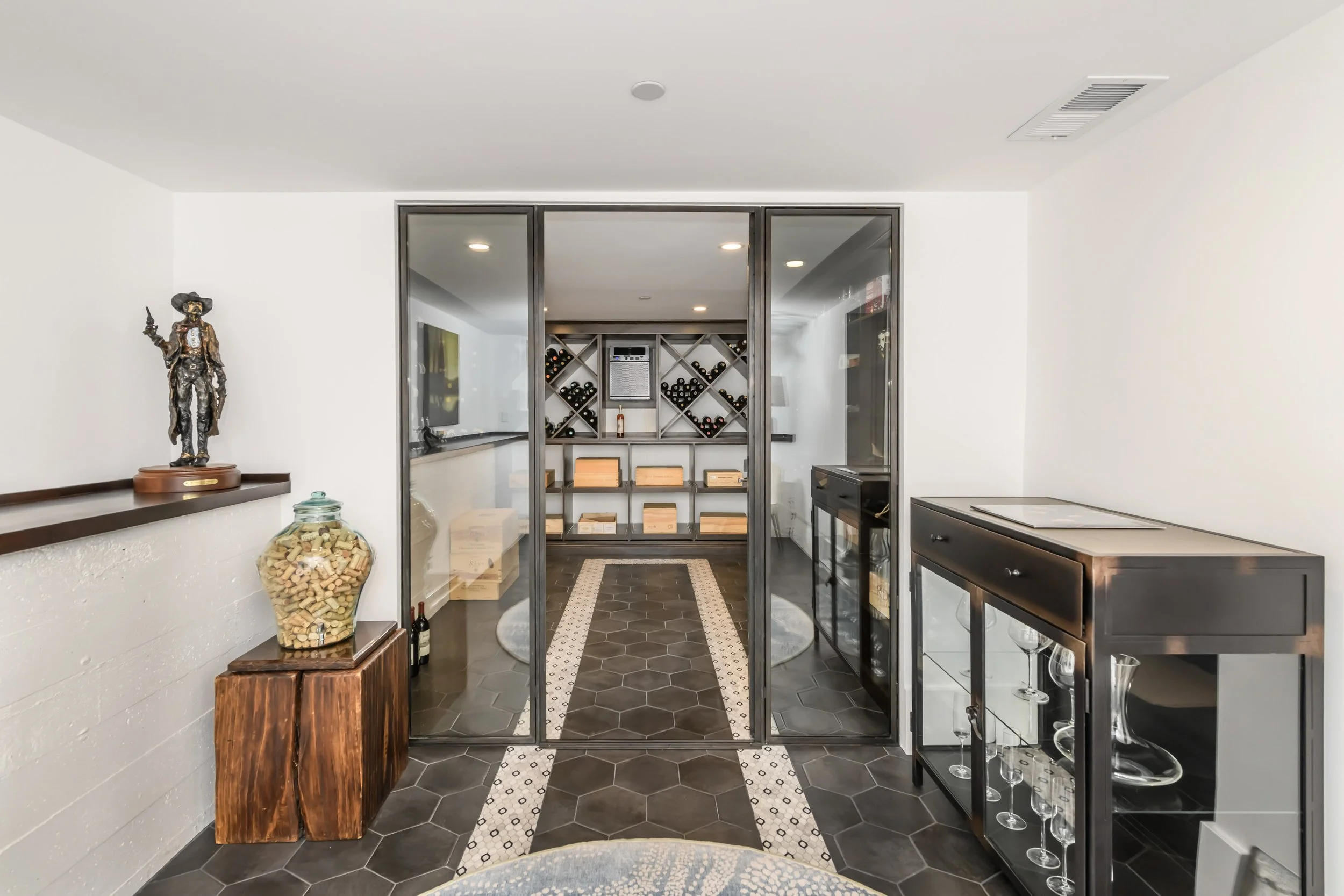 Interior view of a stylish home wine cellar with glass doors, wine racks, and a tiled floor, featuring a decorative statue on a wooden side table and a large jar filled with corks.